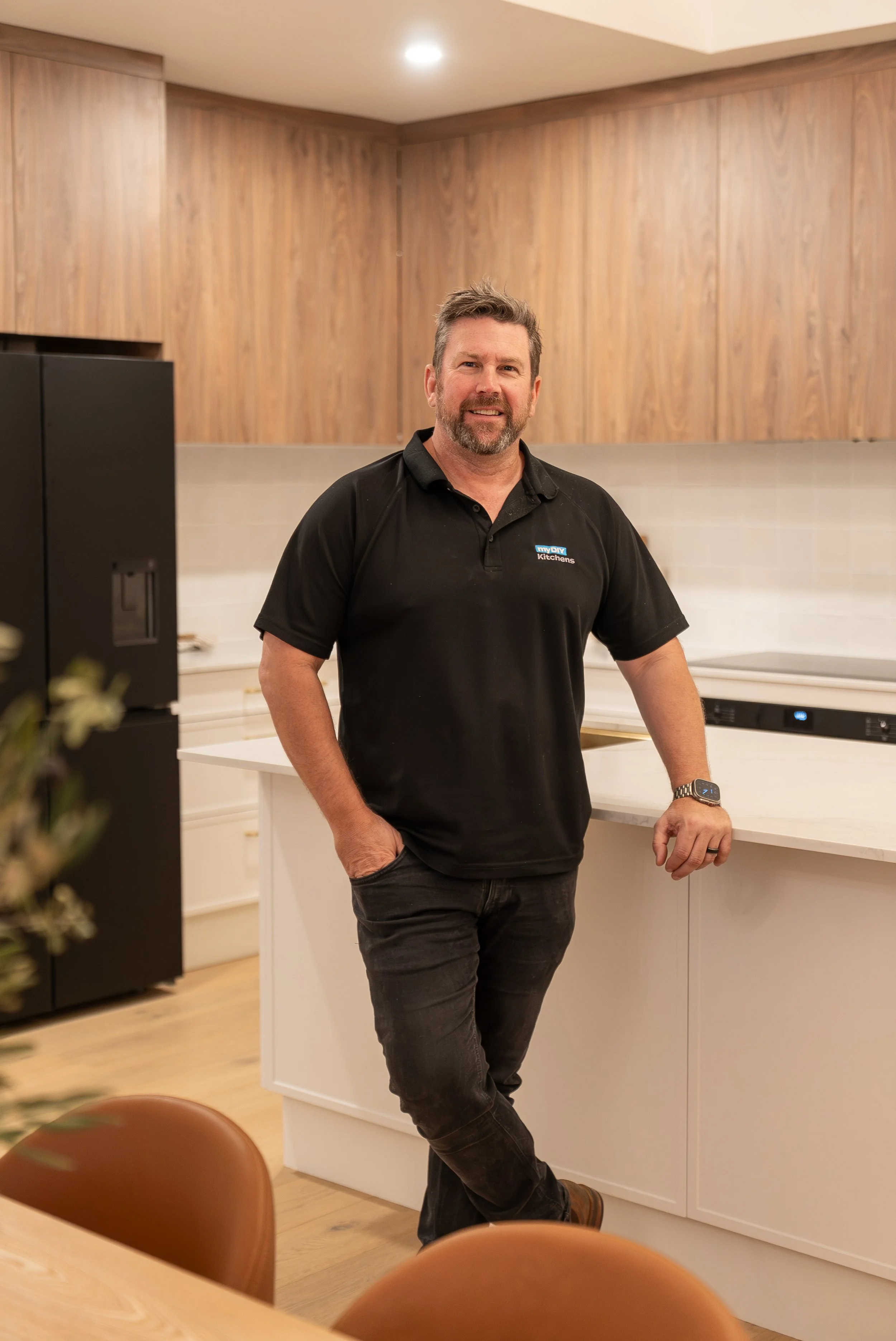 A man with short hair and a beard is standing in a modern kitchen, leaning against the white countertop with one hand in his pocket. He is wearing a black polo shirt with a logo on the chest, black jeans, and a smartwatch. The kitchen features wooden cabinets, a black refrigerator, and a light-colored countertop.