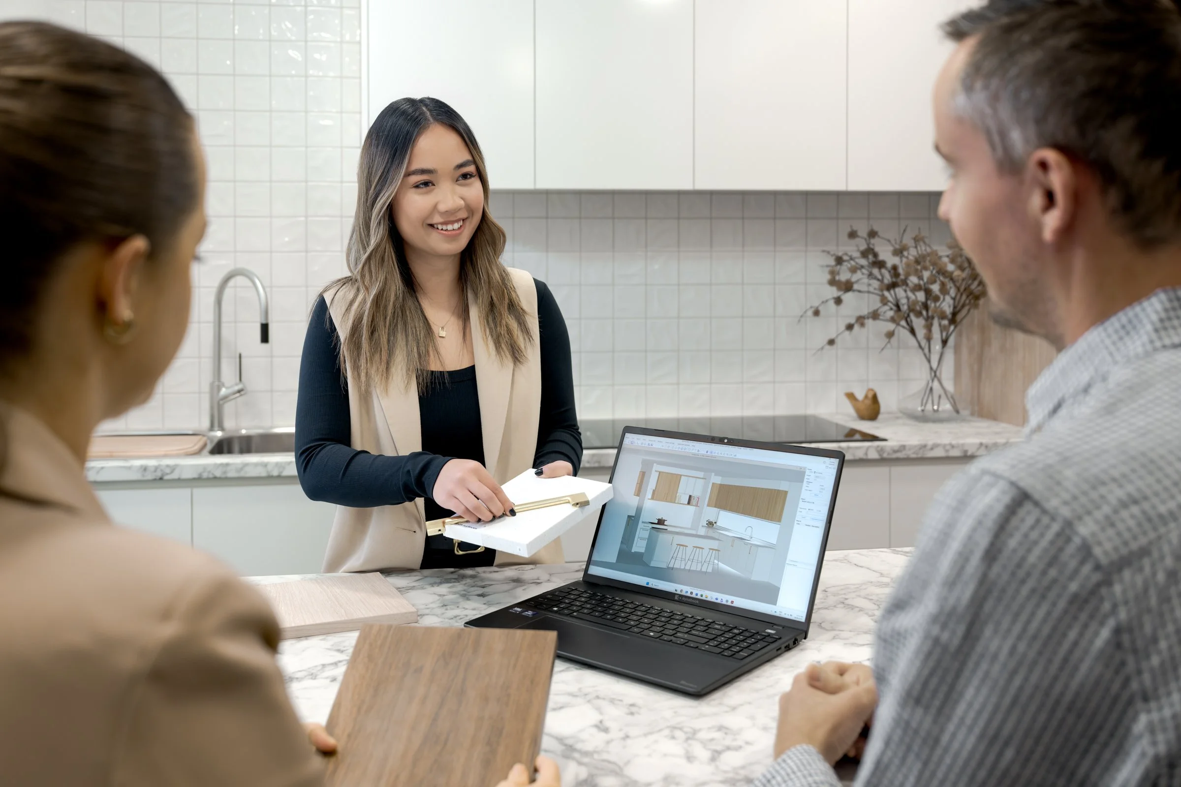 Three people in a kitchen, one woman standing and explaining, two seated with a laptop displaying a kitchen design, during a discussion.