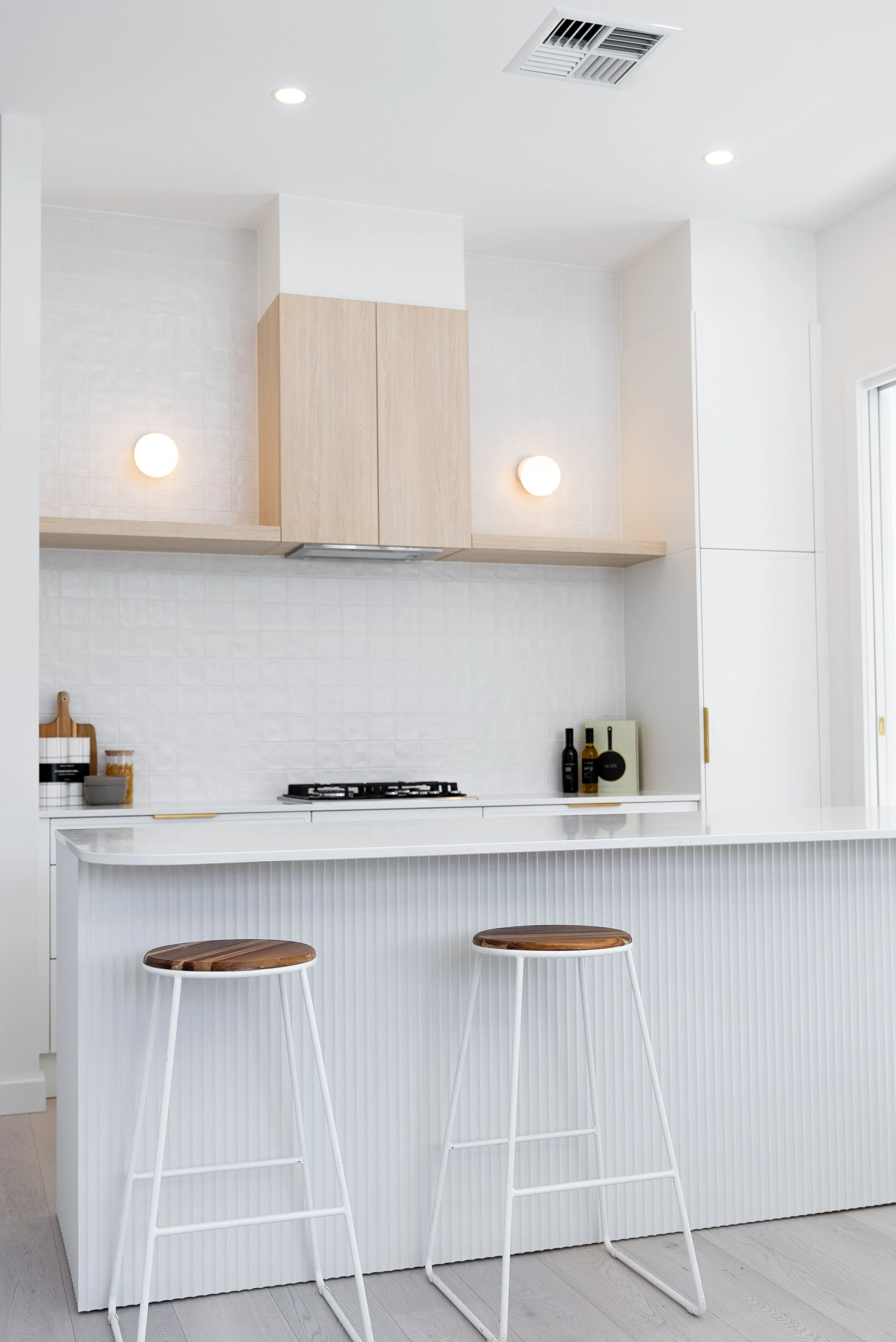 Modern minimalist kitchen with white cabinetry, a white textured island with two wooden stools, light wood upper cabinets, and soft lighting.