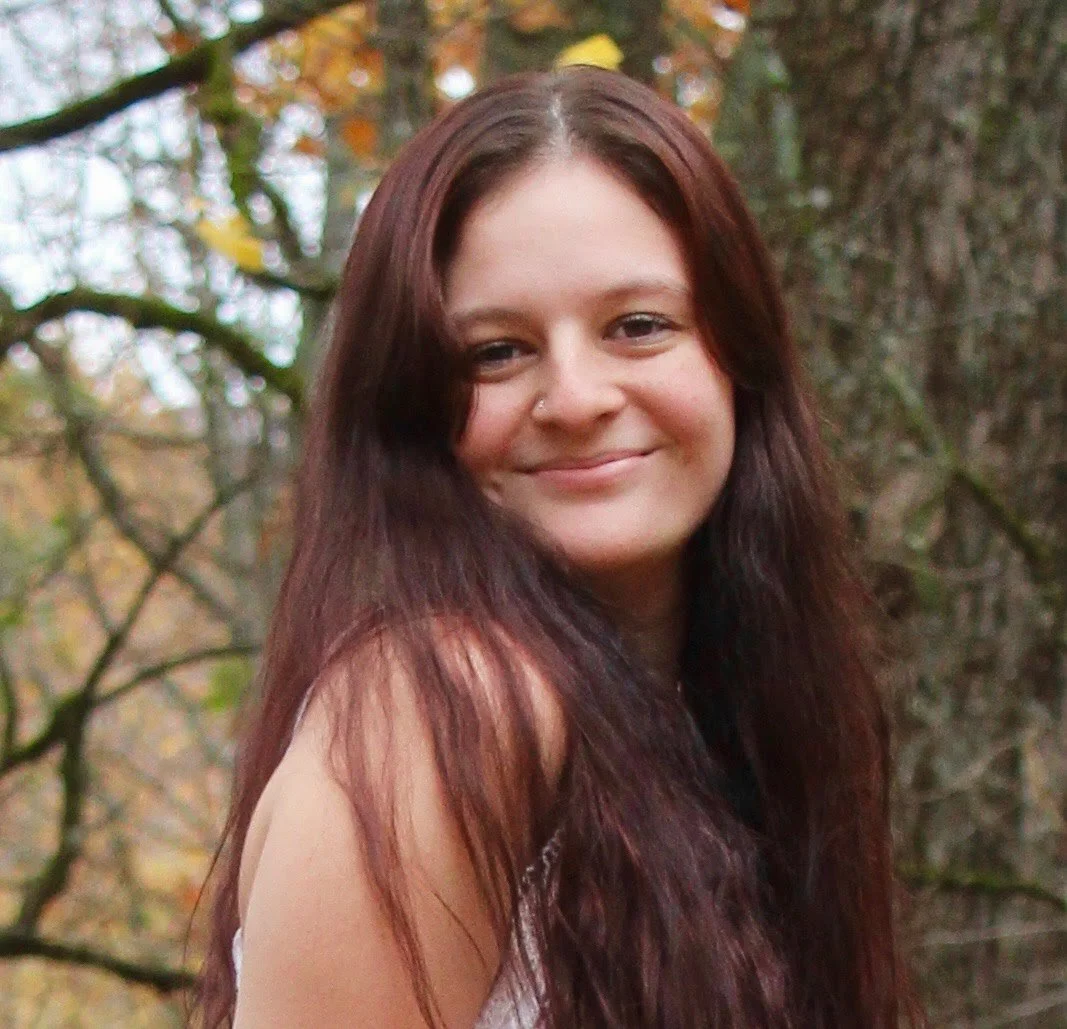 A woman with long, dark brown hair and fair skin smiling outdoors, with trees and branches in the background.