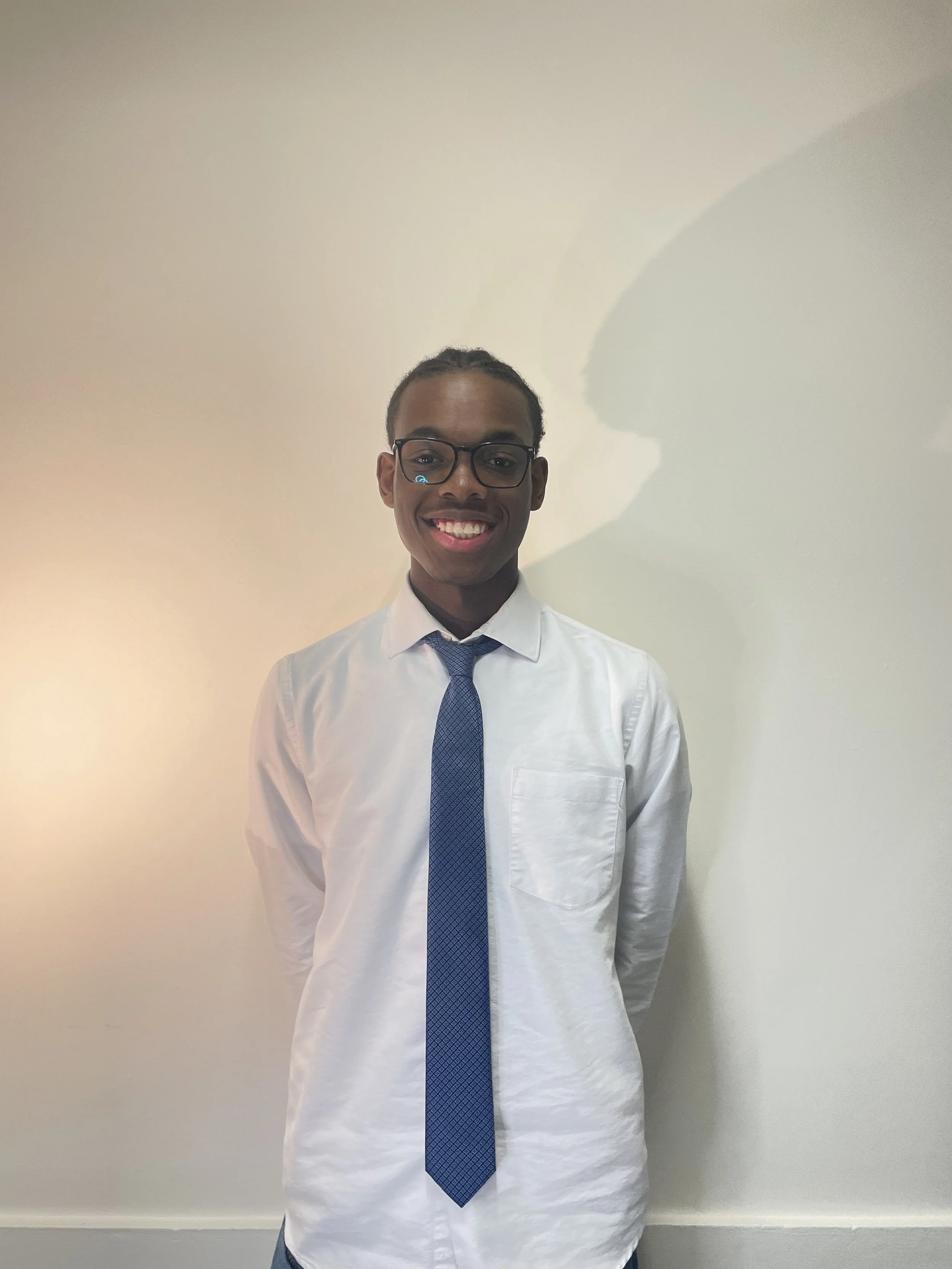Un jeune homme souriant, portant une chemise blanche, une cravate bleue et des lunettes, posé devant un mur blanc.