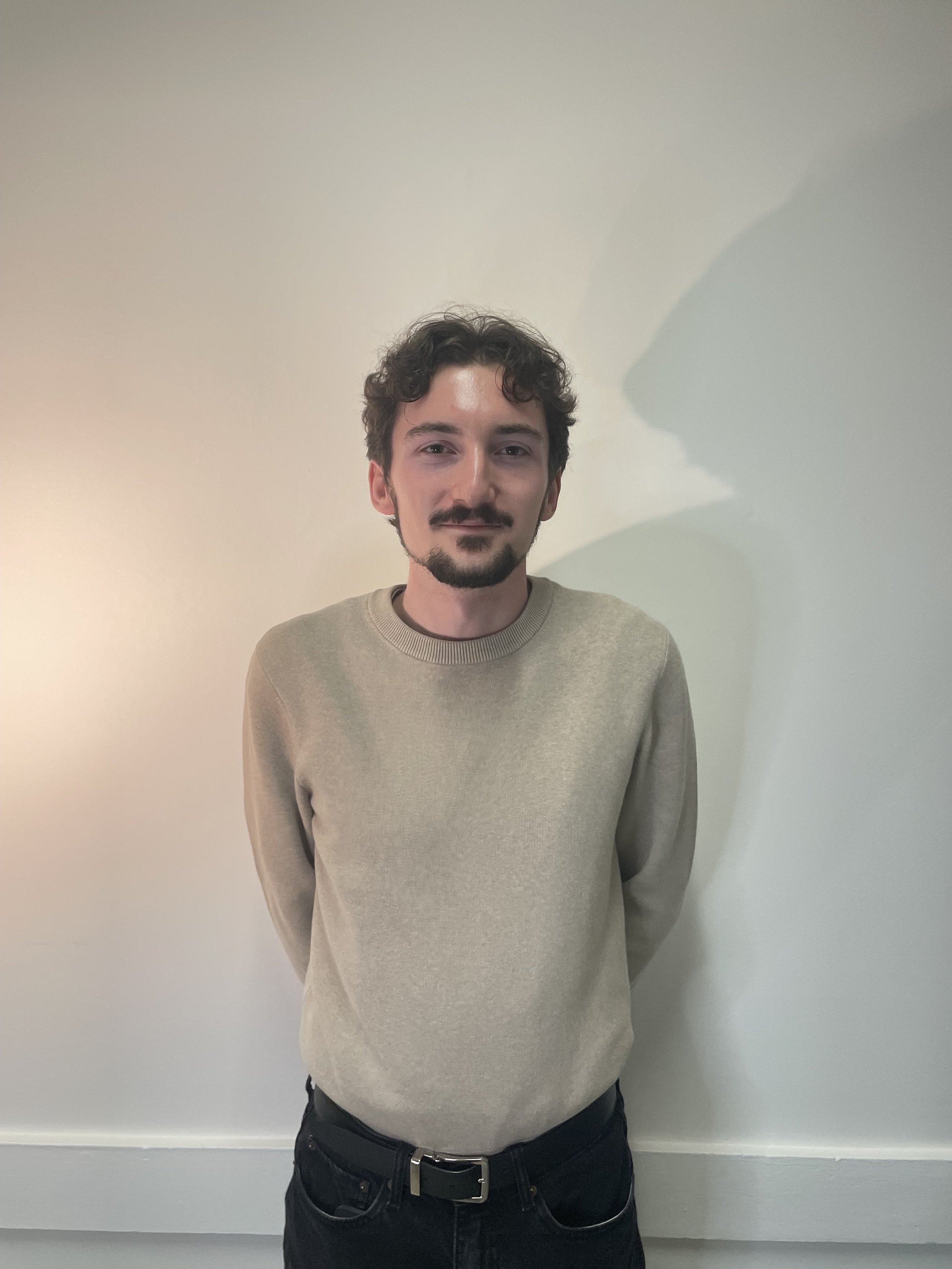 Un jeune homme avec des cheveux bouclés et une barbe, portant un sweat beige, se tient devant un mur clair.