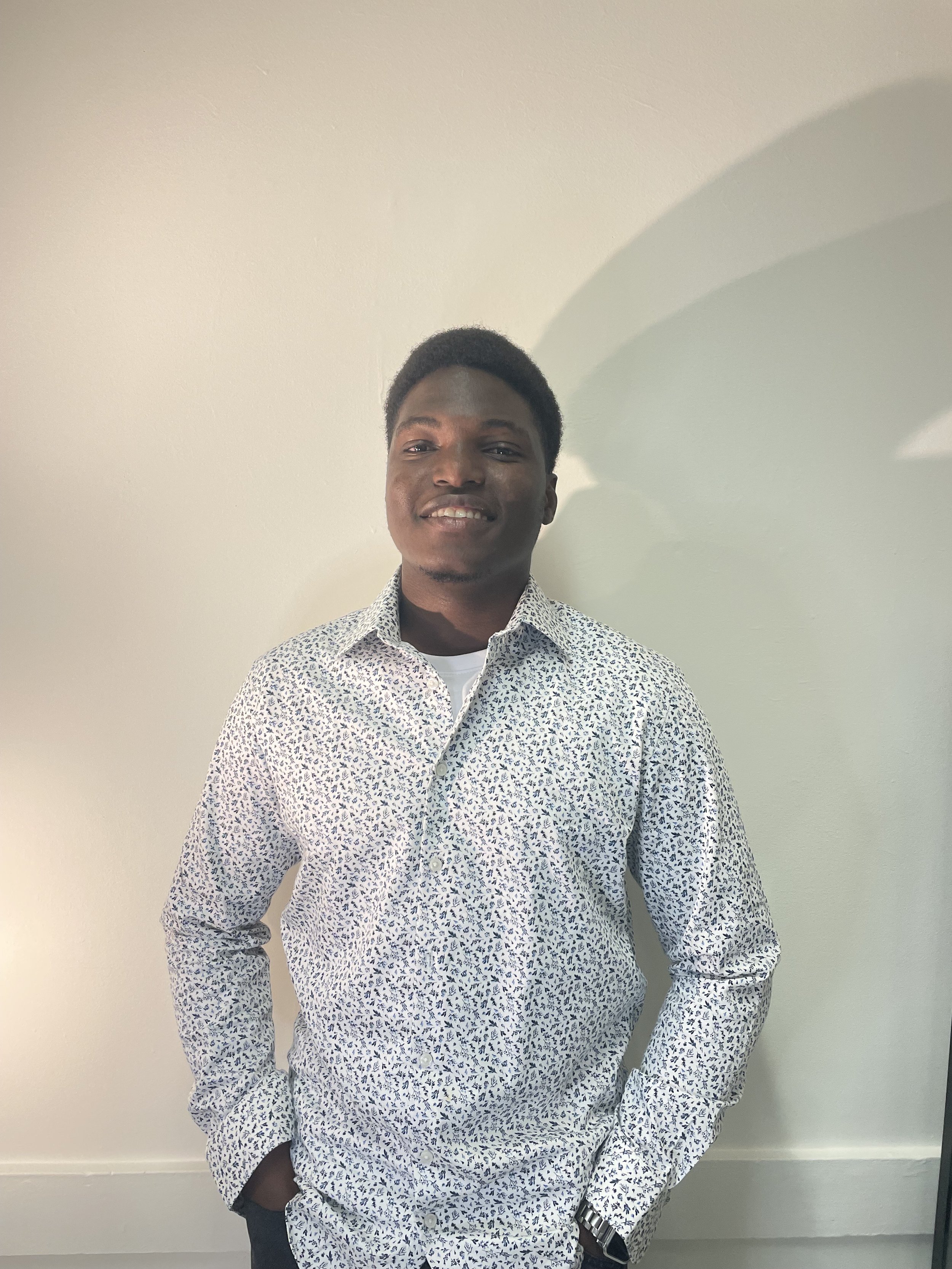 Un jeune homme souriant portant une chemise à motifs blancs et bleus, posant contre un mur blanc.