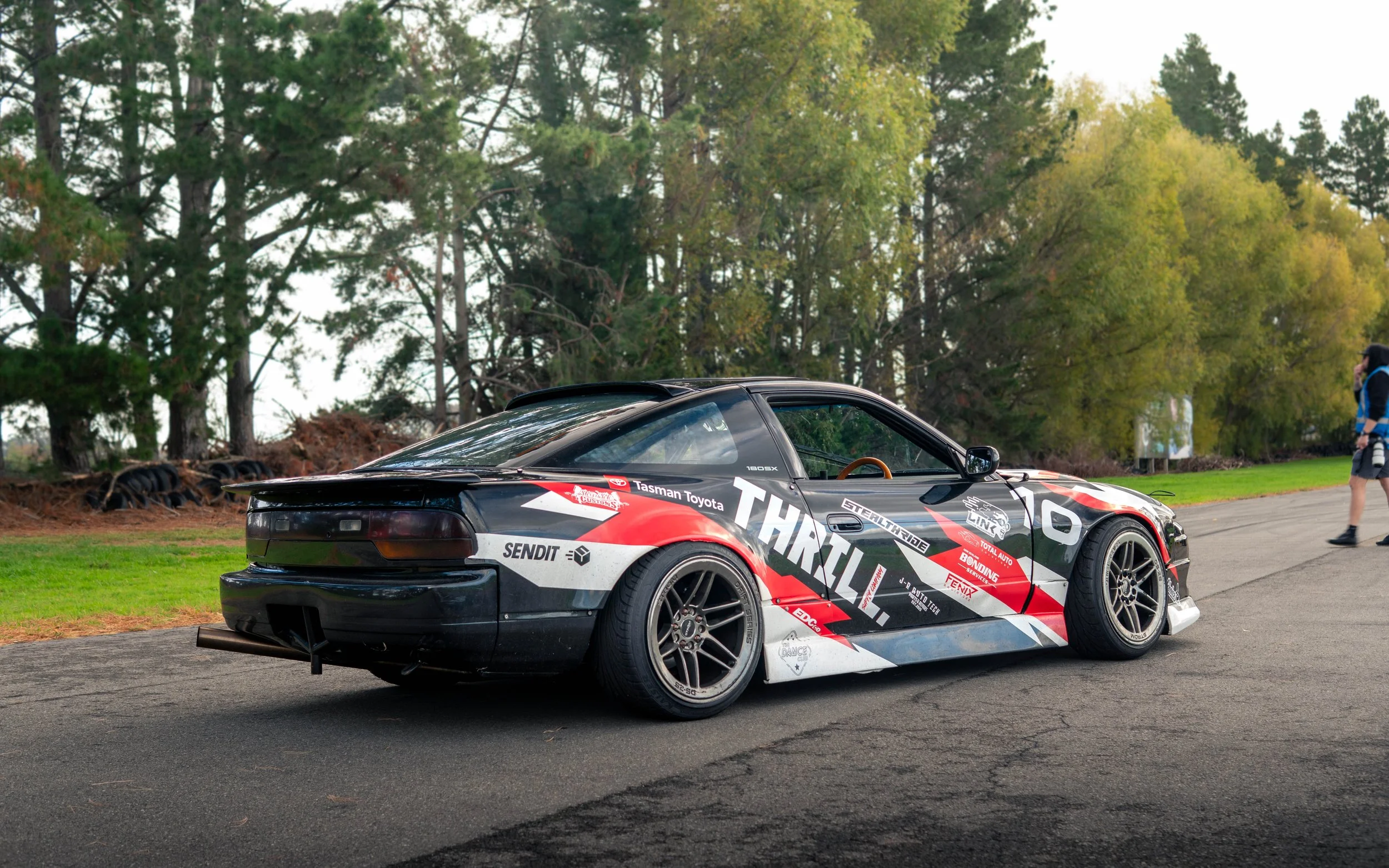 A sports car with a black, red, and white racing design parked on a paved surface near a grassy area and surrounded by tall trees. A person holding a camera walks near the car.