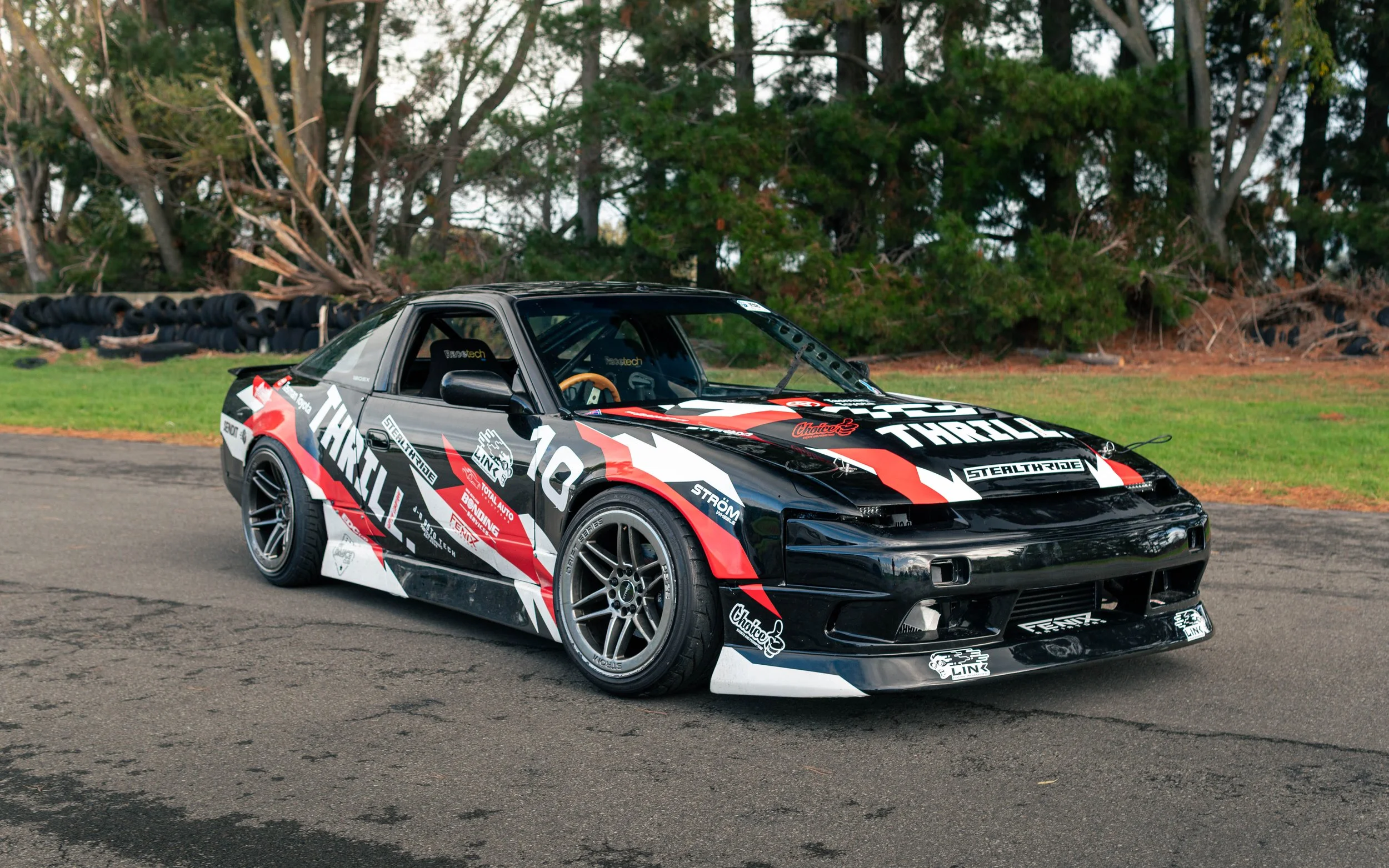 A black, red, and white race car with various sponsor logos parked on a paved surface with tire stacks and green trees in the background.