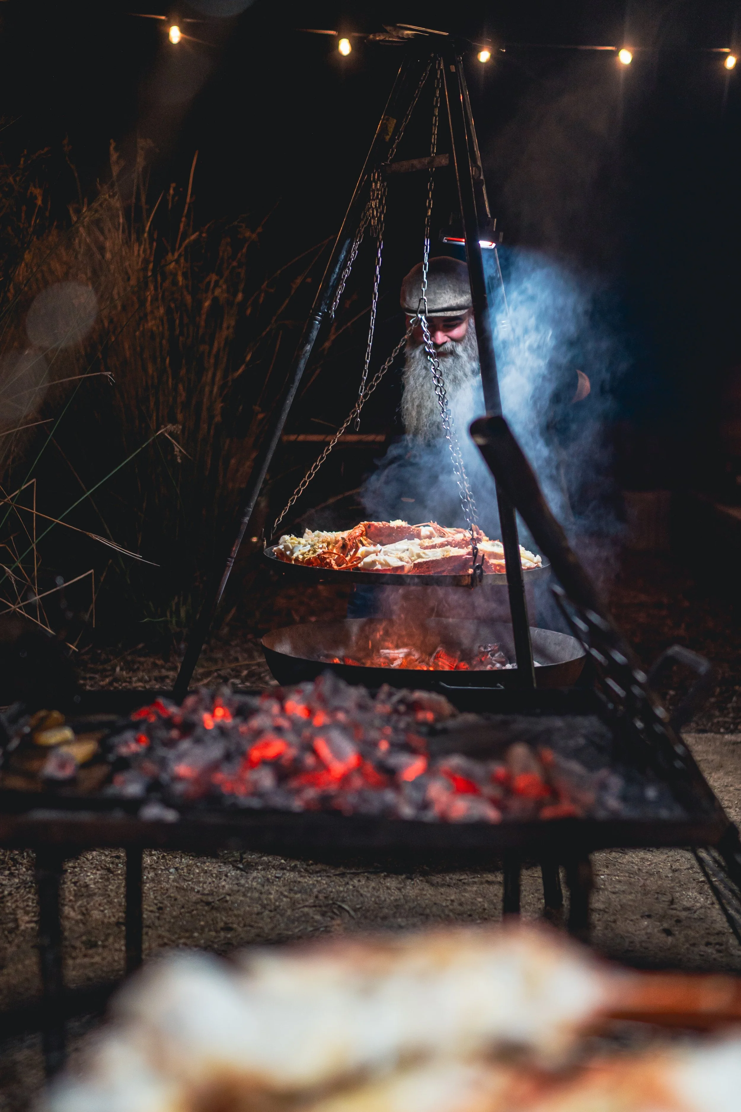 Tasting Trail's TrailGraze Feast | A man with a grey beard and cap cooking food over an open fire outdoors at night, with string lights overhead.