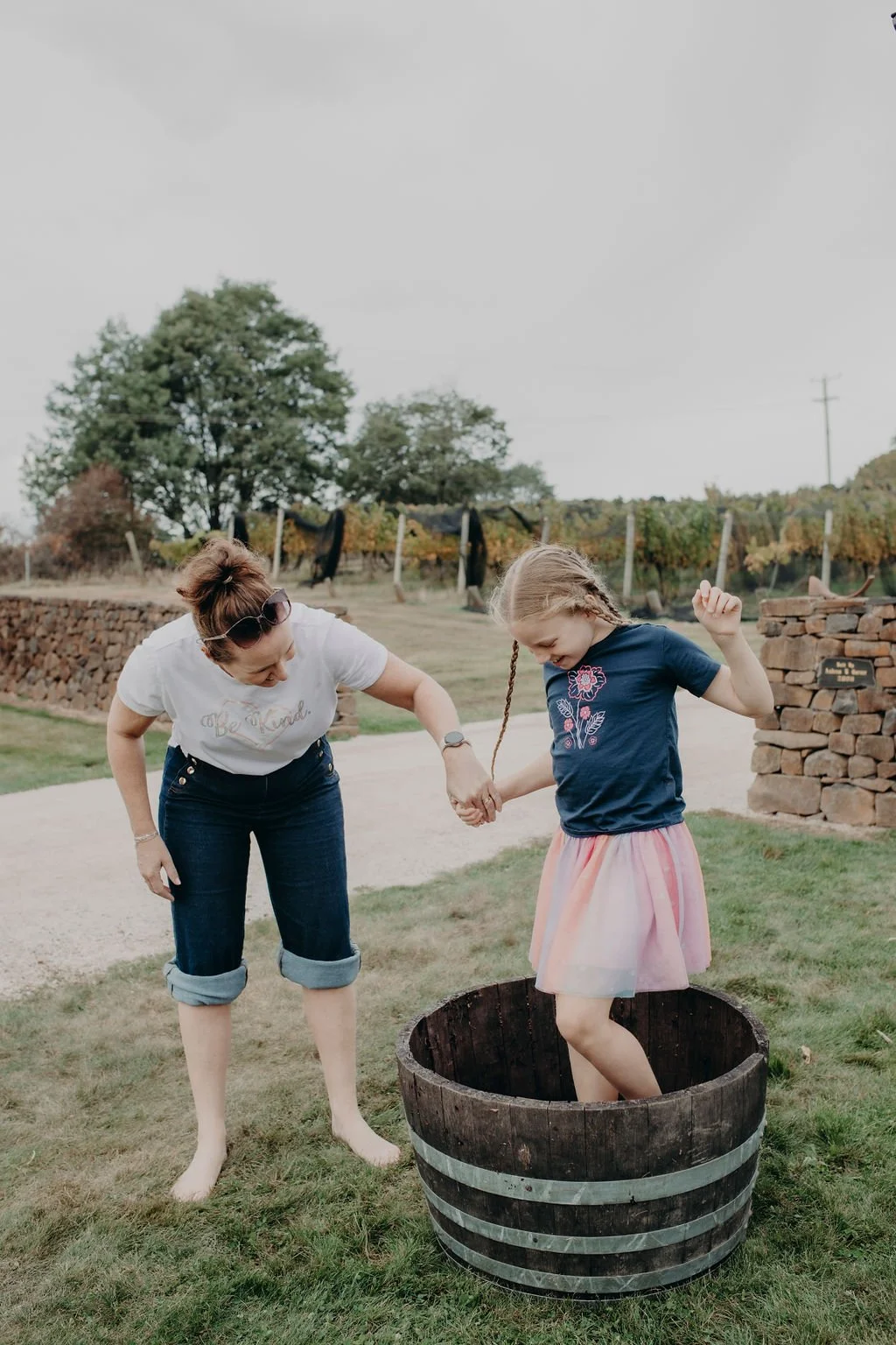 A woman helping a young girl out of a wine barrel, holding hands outdoors on a cloudy day.