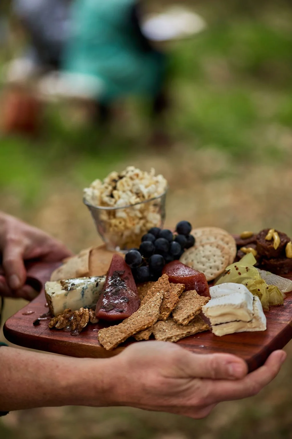 Tasting Trail Tasmania | Person holding a wooden cheese and charcuterie board with cheese, crackers, grapes, and cured meats, with a bowl of popcorn in the background and a blurred outdoor setting.