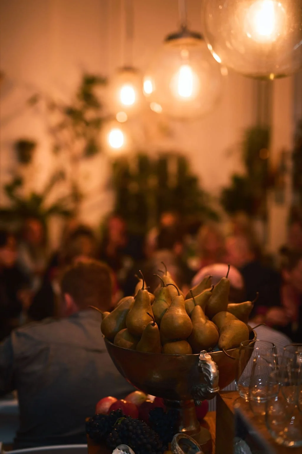 A bowl of pears on a table at a dimly lit gathering or dinner party, with people and hanging lights in the background.