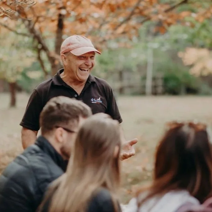 An older man smiling and talking to a group of young people outdoors in a park during fall with orange leaves on trees.