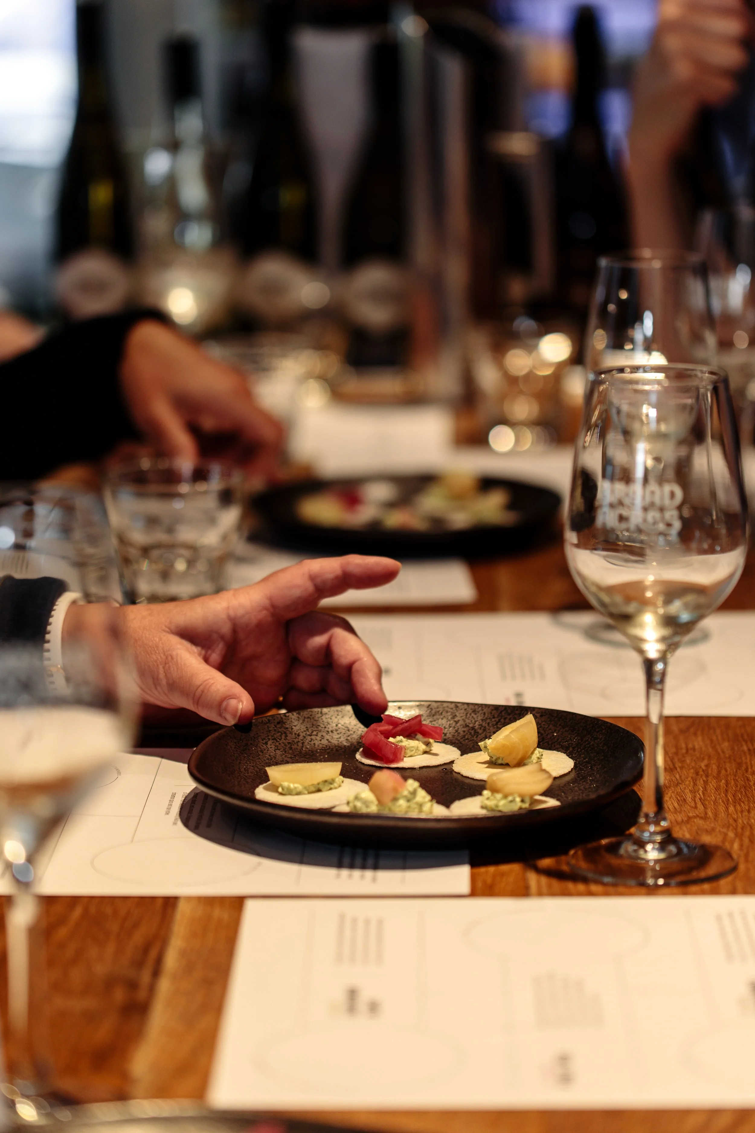 A person is selecting a small appetizer from a black plate at a dining table, surrounded by wine glasses and paper menus, with a relaxed dining ambiance.