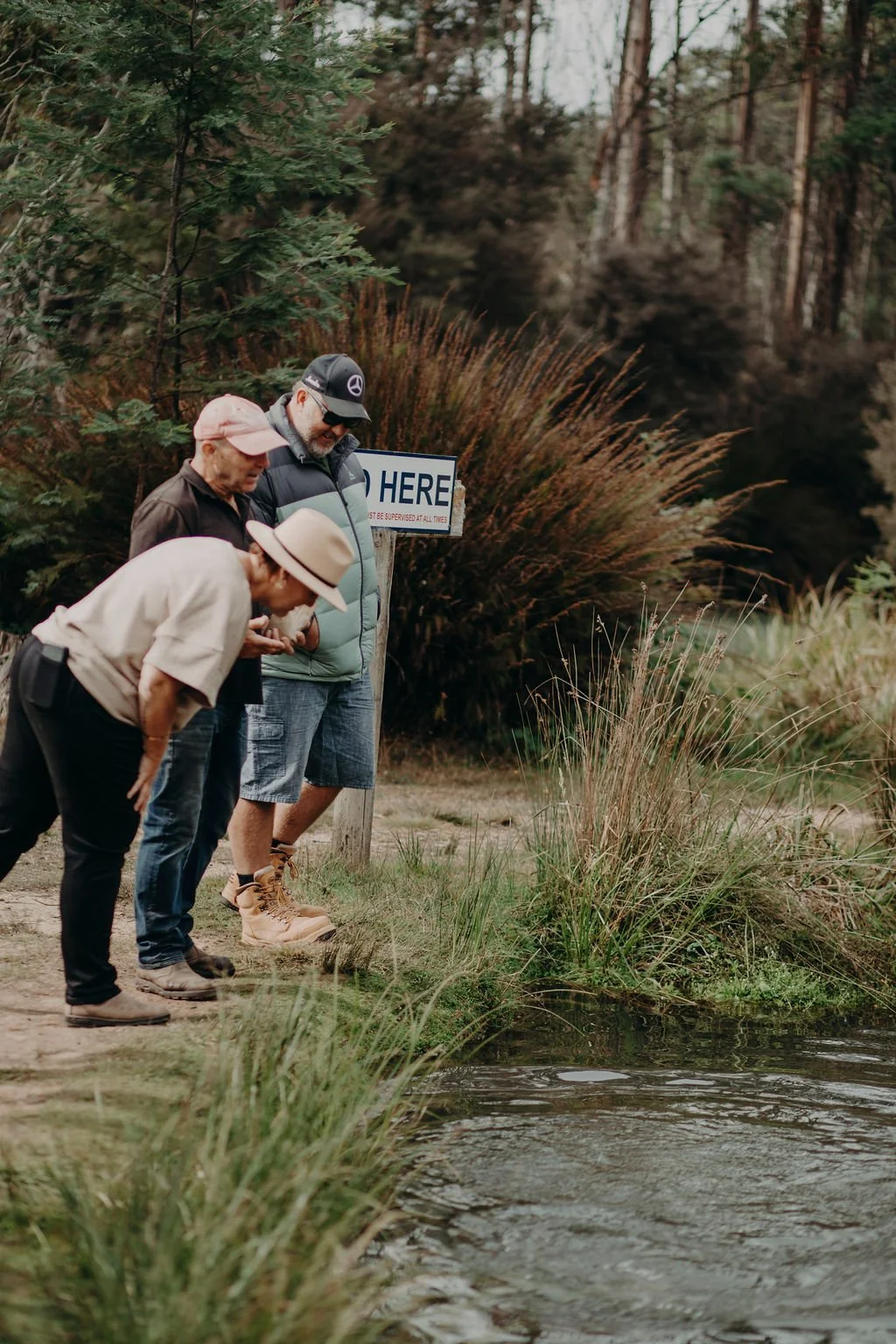 Three people, two women and one man, are standing at the edge of a small pond in a wooded area, looking into the water. They are dressed casually for outdoor activity, and a sign that says 'NO FISHING HERE' is visible in the background.