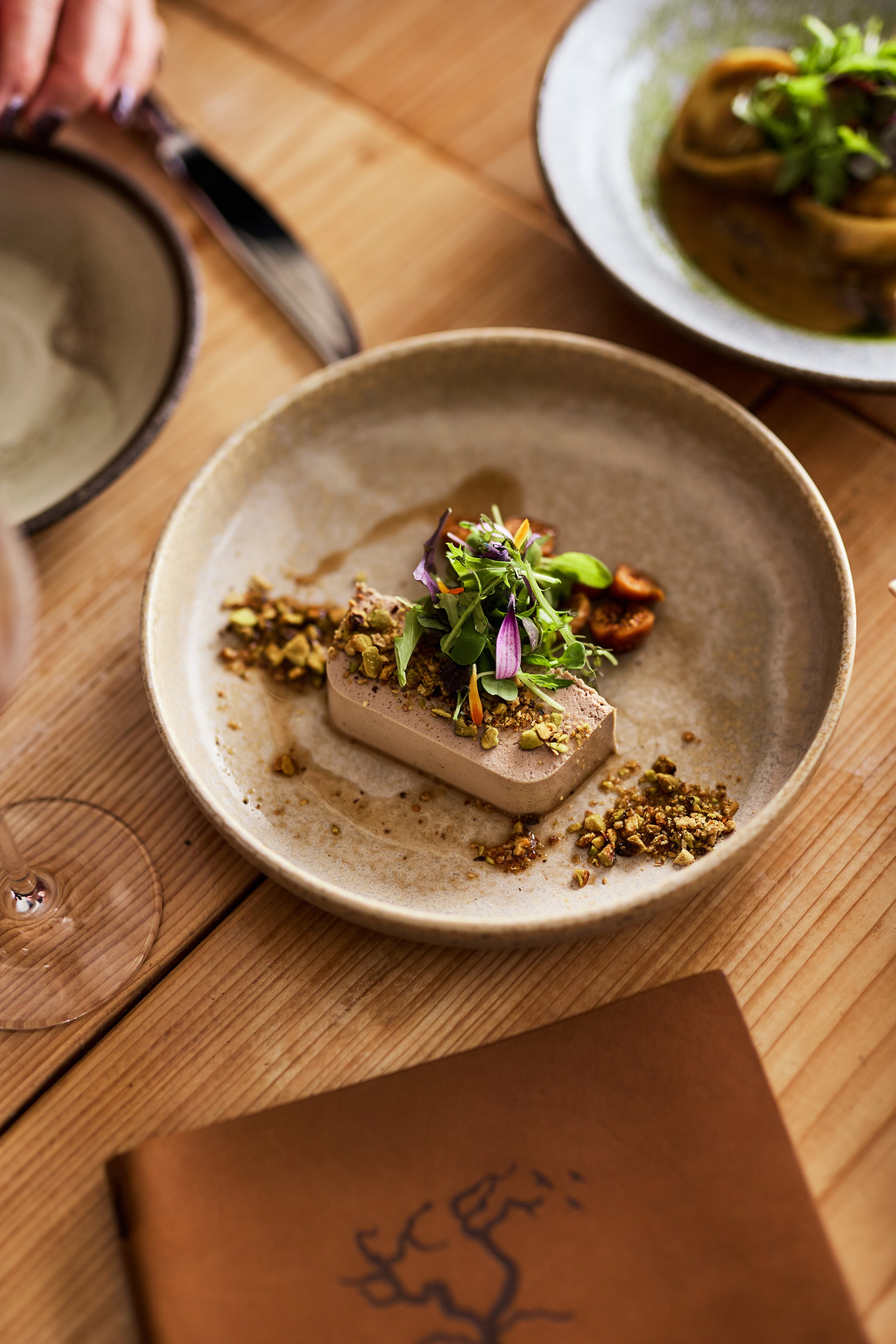 Tasting Trail Tasmania | A plated dessert topped with microgreens and crushed pistachios, served on a ceramic plate on a wooden table with a fork, wine glass, and another dish in the background.