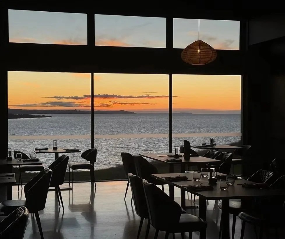 Interior of Basalt restaurant with large windows showing a sunset over the ocean, tables set with glassware, and chairs arranged for dining.