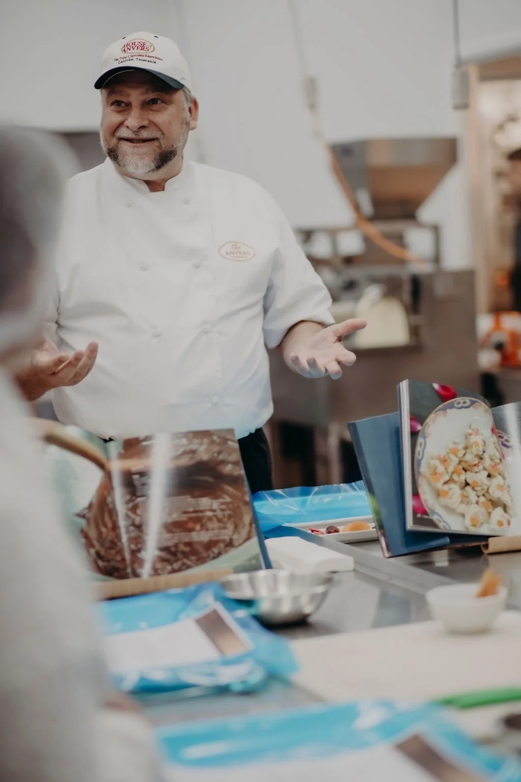 Anvers Chocolate's Chocolatier Igor in a white uniform and cap is talking to a person as they learn how to make chocolate at a TrailGraze chocolate making session.