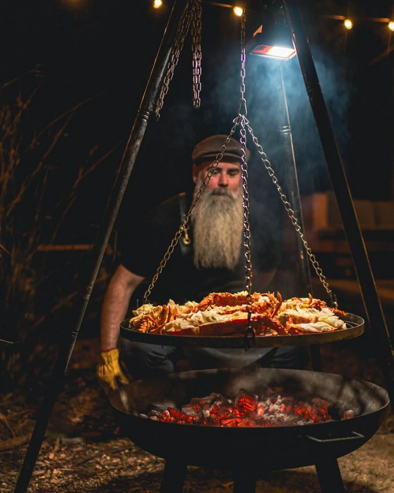 TrailGraze Feast | A man with a beard wearing a cap and gloves cooking a large pile of food on a hanging grill over an open fire at night, with string lights overhead.
