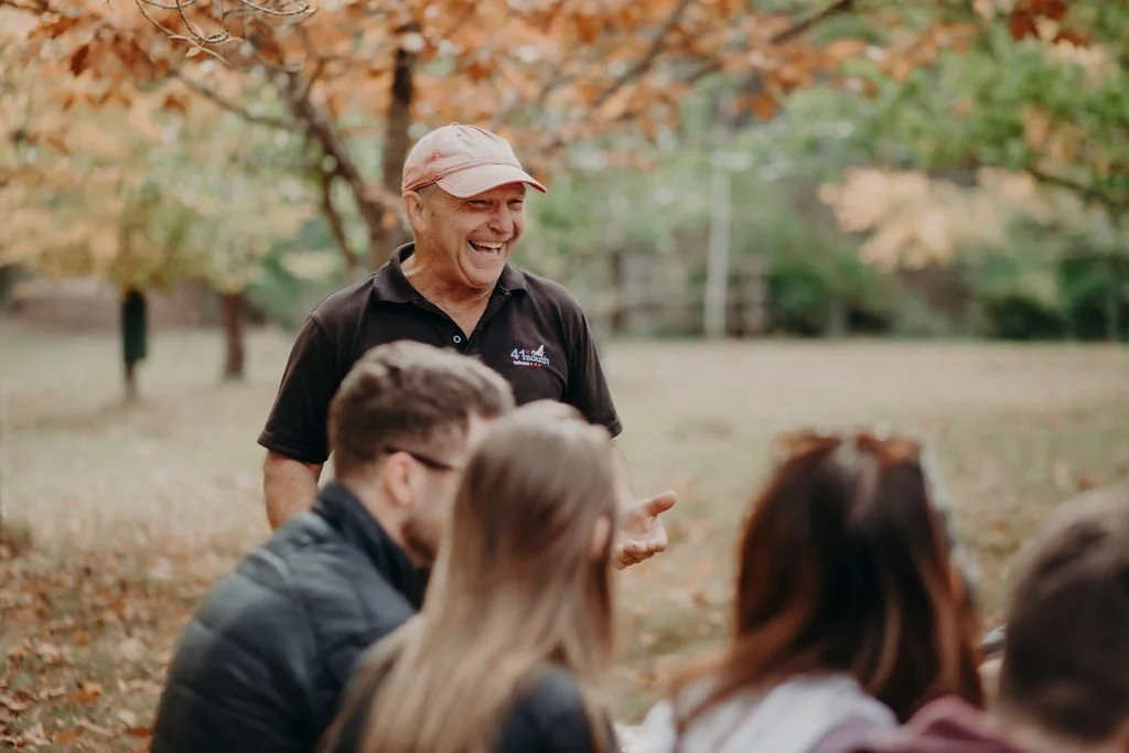 Tasting Trail Tasmania | An elderly man with a beige cap and black shirt smiling and talking to a group of people outdoors during autumn, with trees and fallen leaves in the background.