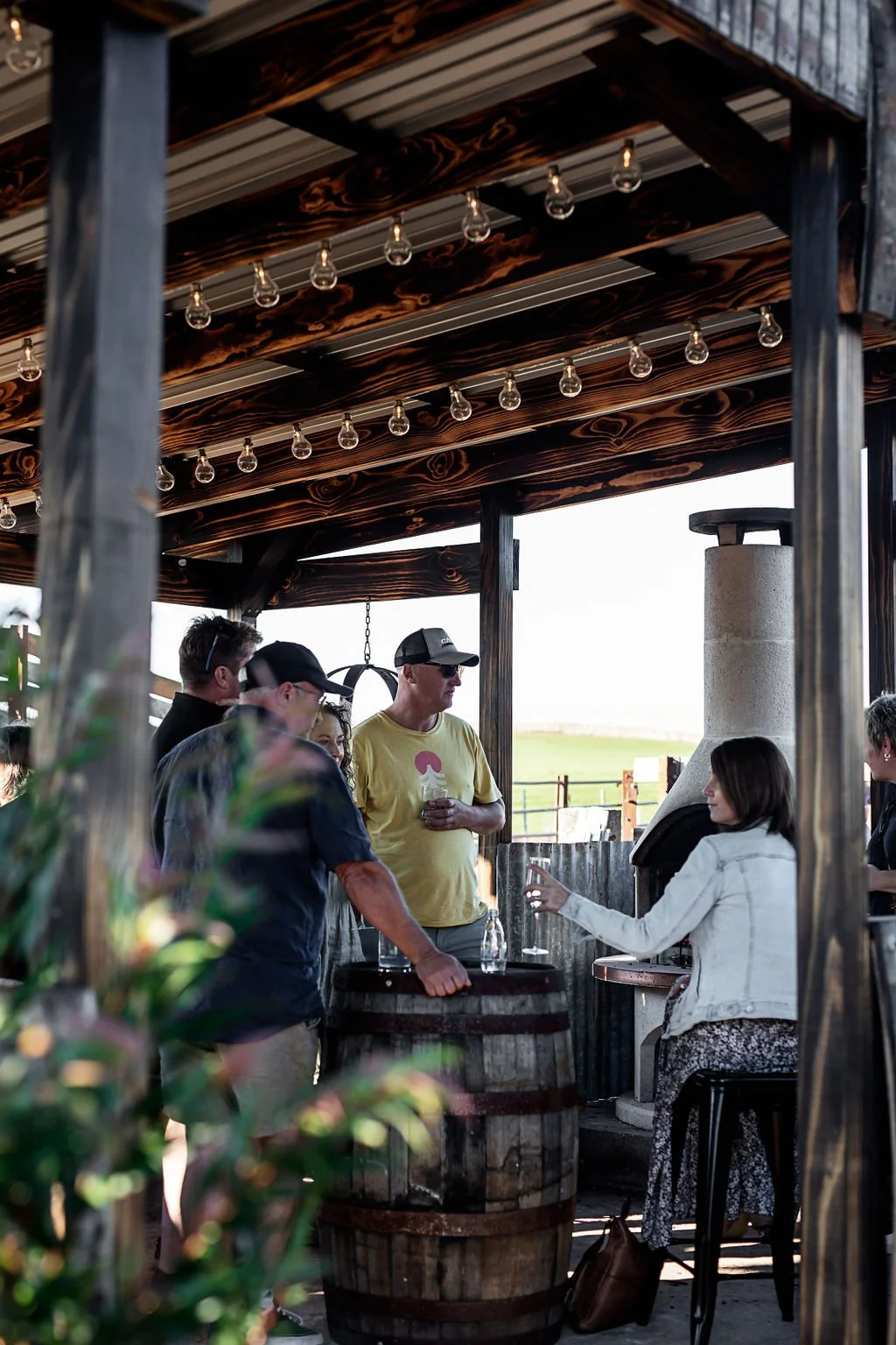 TrailGraze | Group of people socialising at a Alchymia Distillery's rustic outdoor bar with string lights and wooden decor during daytime.