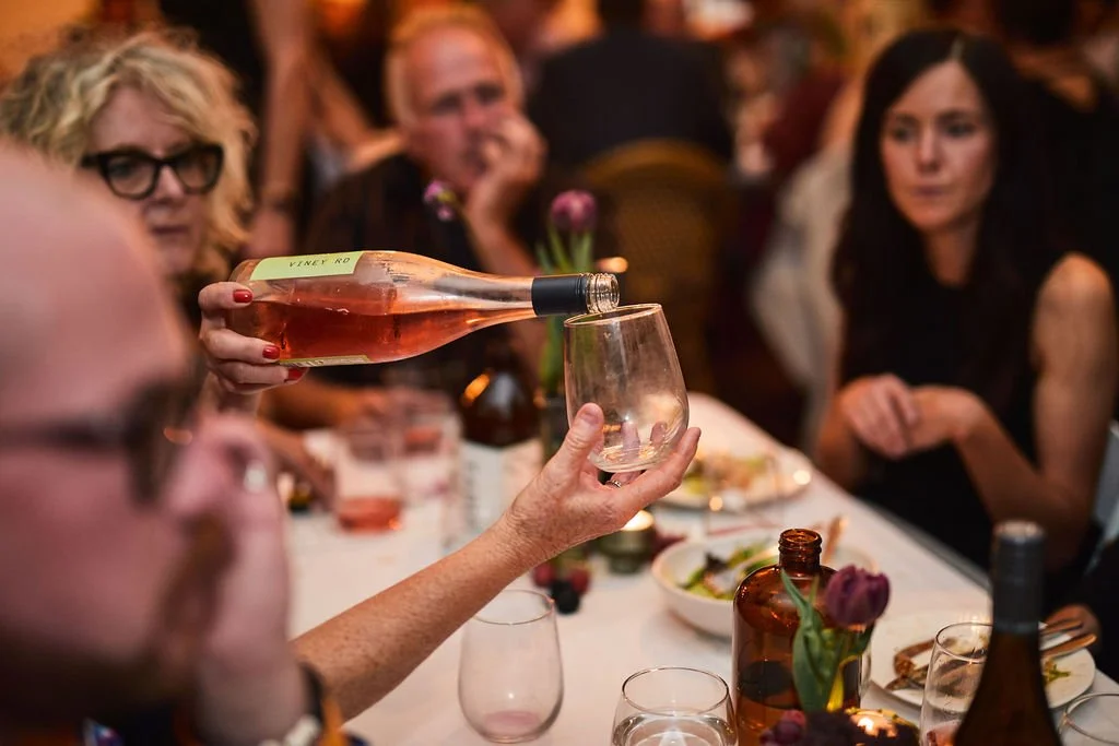 Person pouring rosé wine into a glass at a dinner gathering, with other guests seated around the table.