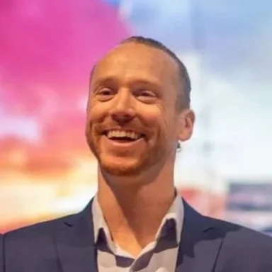 A smiling man in a suit standing in front of a colorful blurred background.