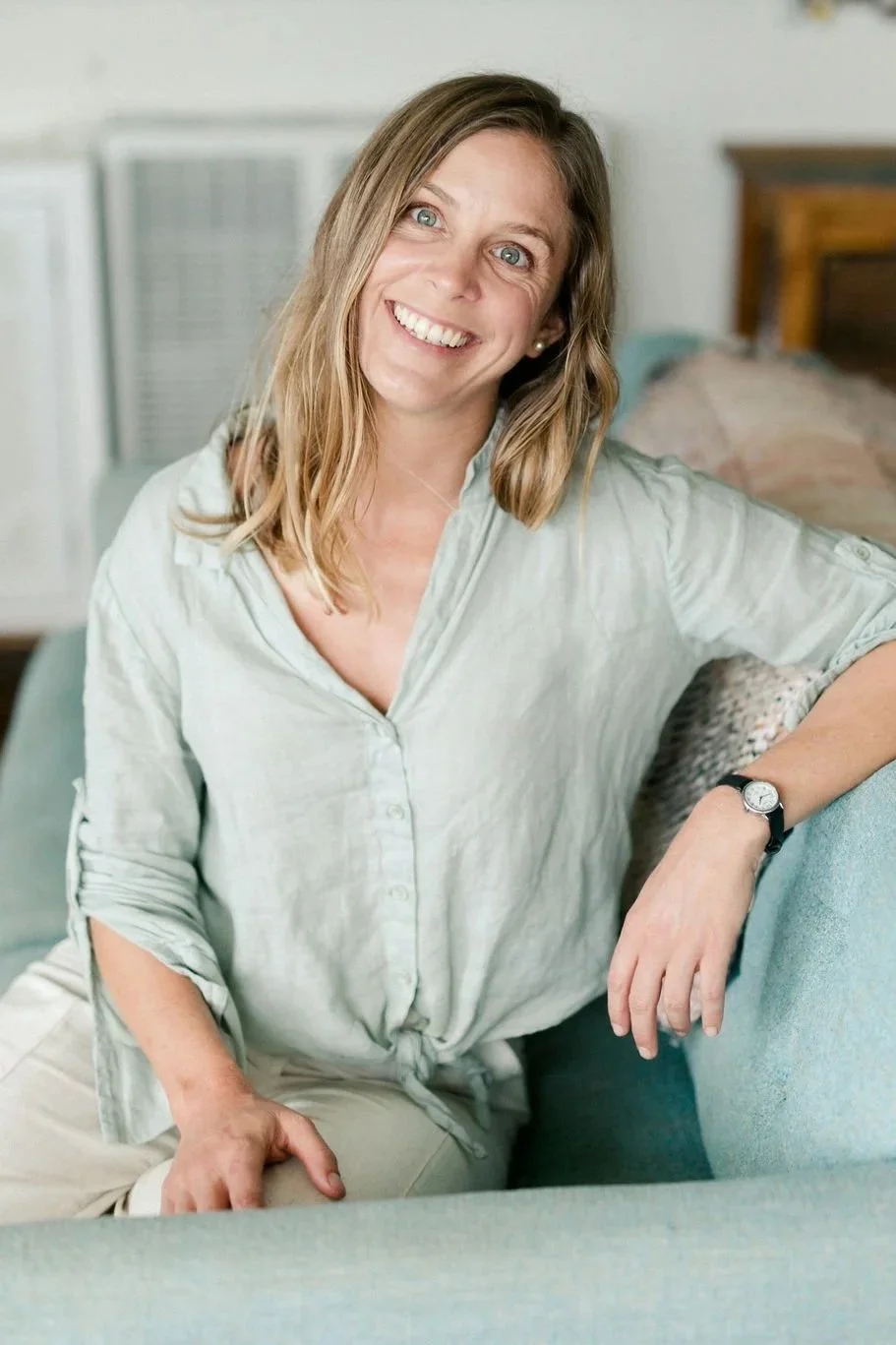 The artist with shoulder-length blonde hair smiling and sitting on a teal sofa in a room, wearing a sage color button-up shirt and a watch.