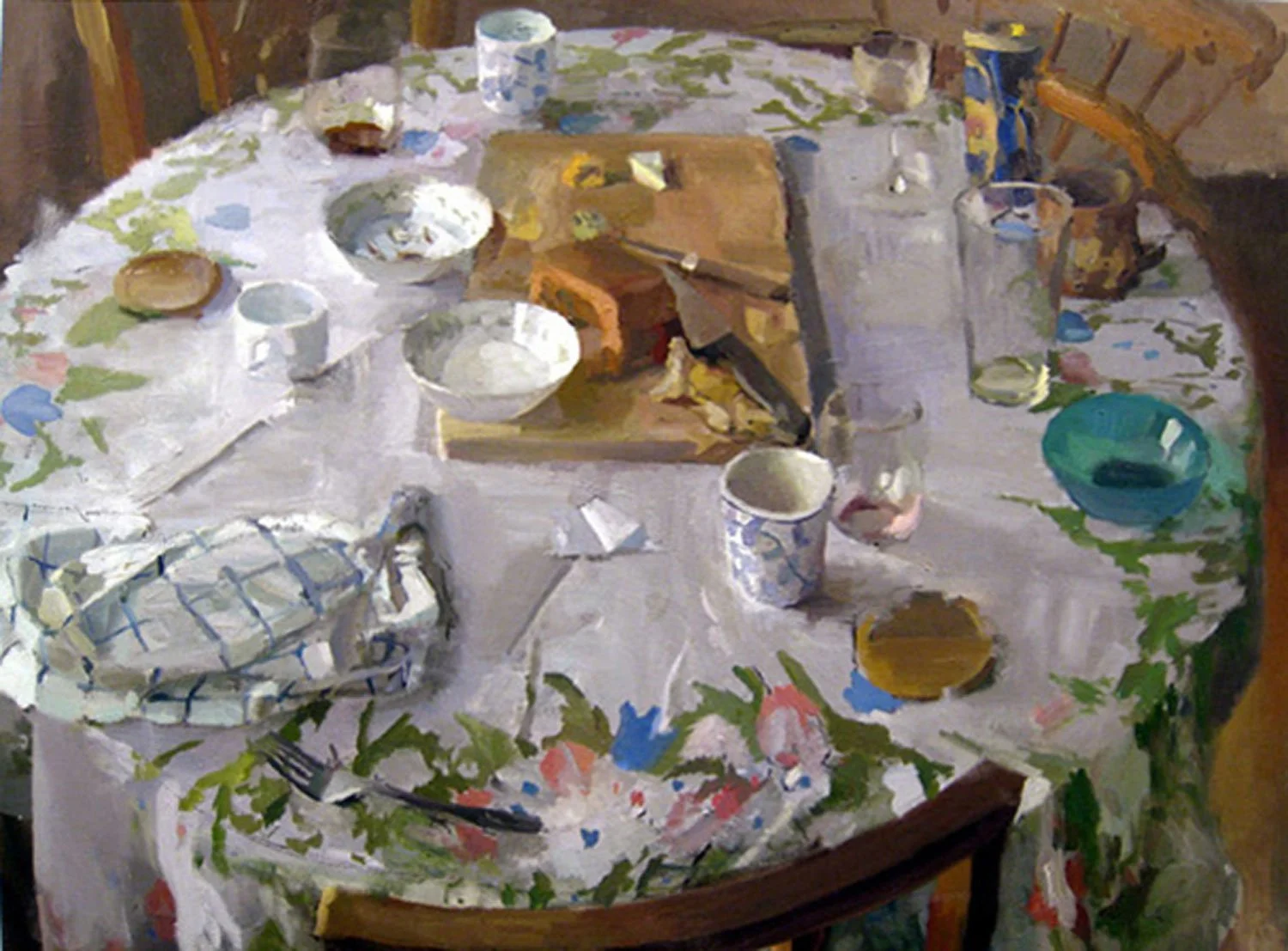 A round dining table covered with a floral tablecloth holds various dishes, cups, bowls, a cutting board, and kitchen items, with chairs around it.