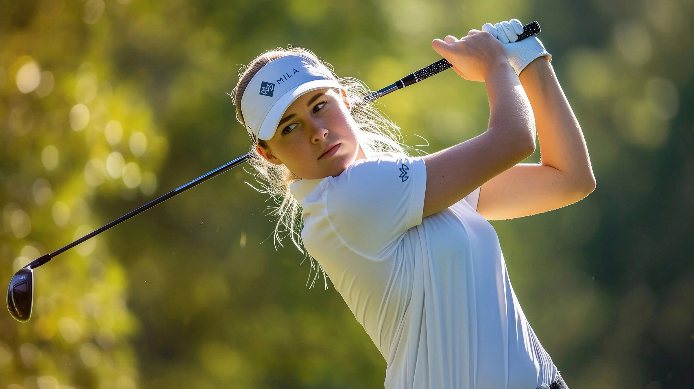 Young woman in white golf outfit swinging a golf club on a golf course with blurred trees in the background.