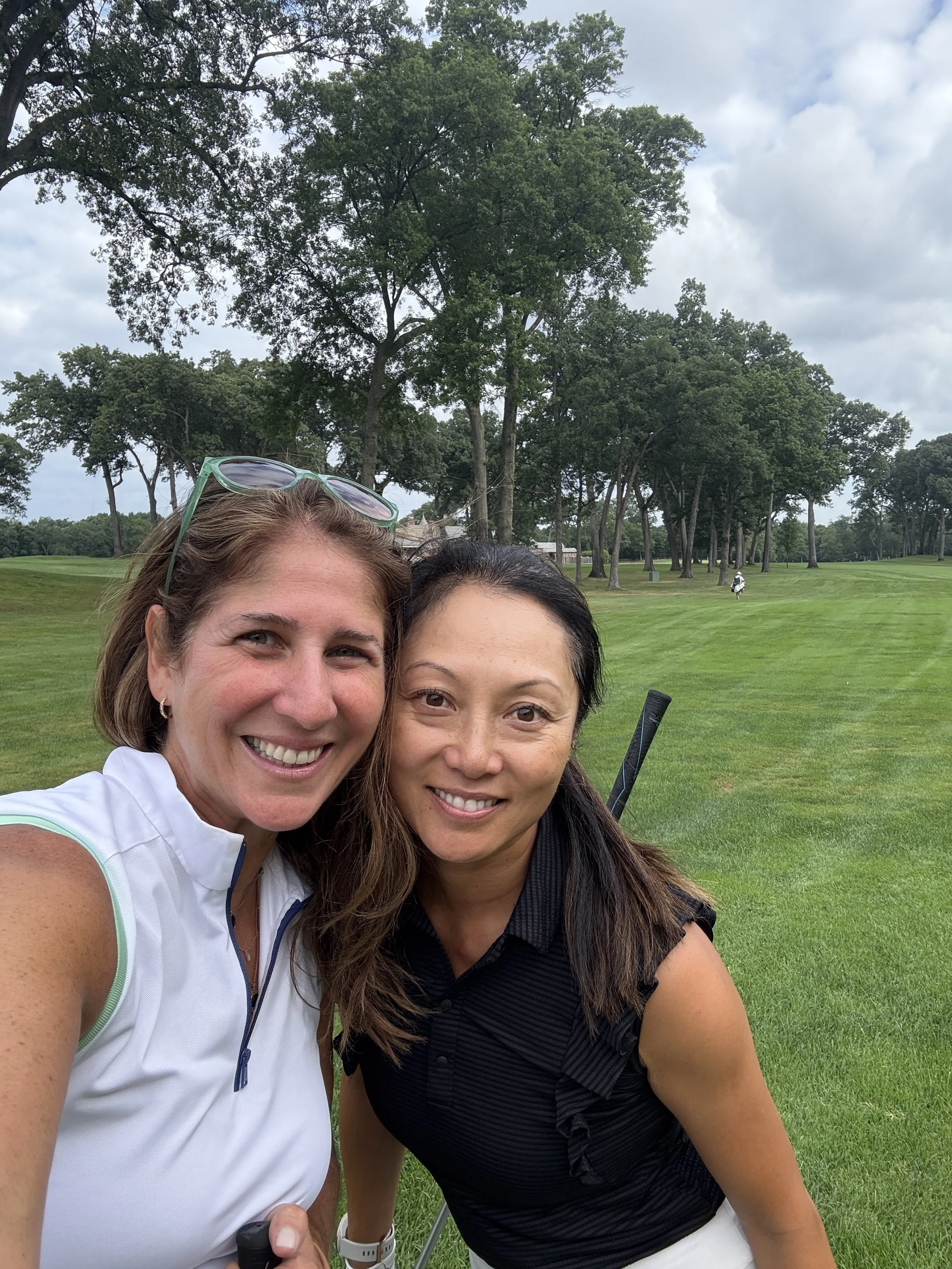 Two women taking a selfie on a golf course, smiling, with trees and a cloudy sky in the background.