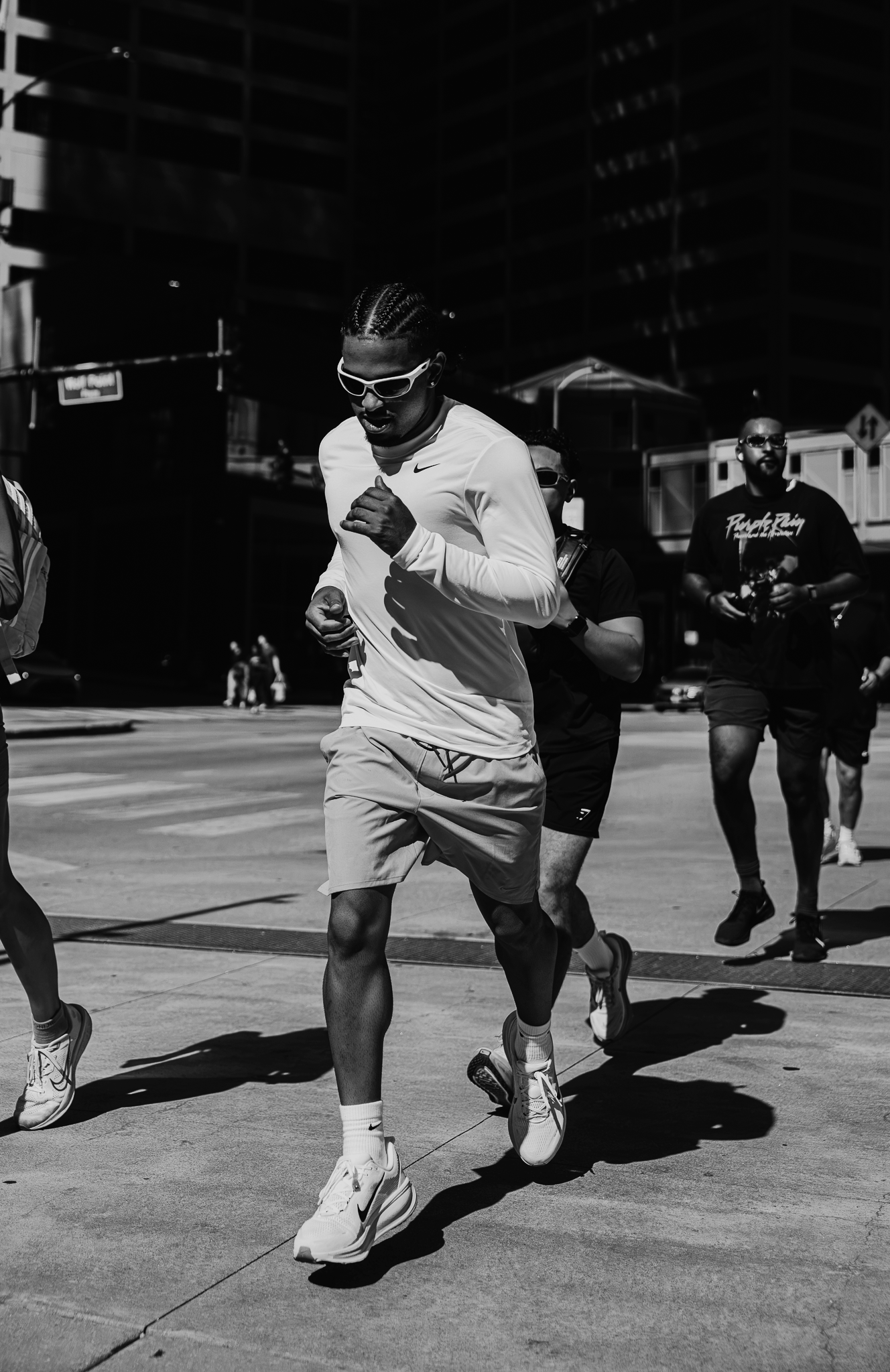 A group of people running outside in an urban area, with tall buildings in the background. The person in the foreground is wearing sunglasses, a light shirt, shorts, and sneakers.