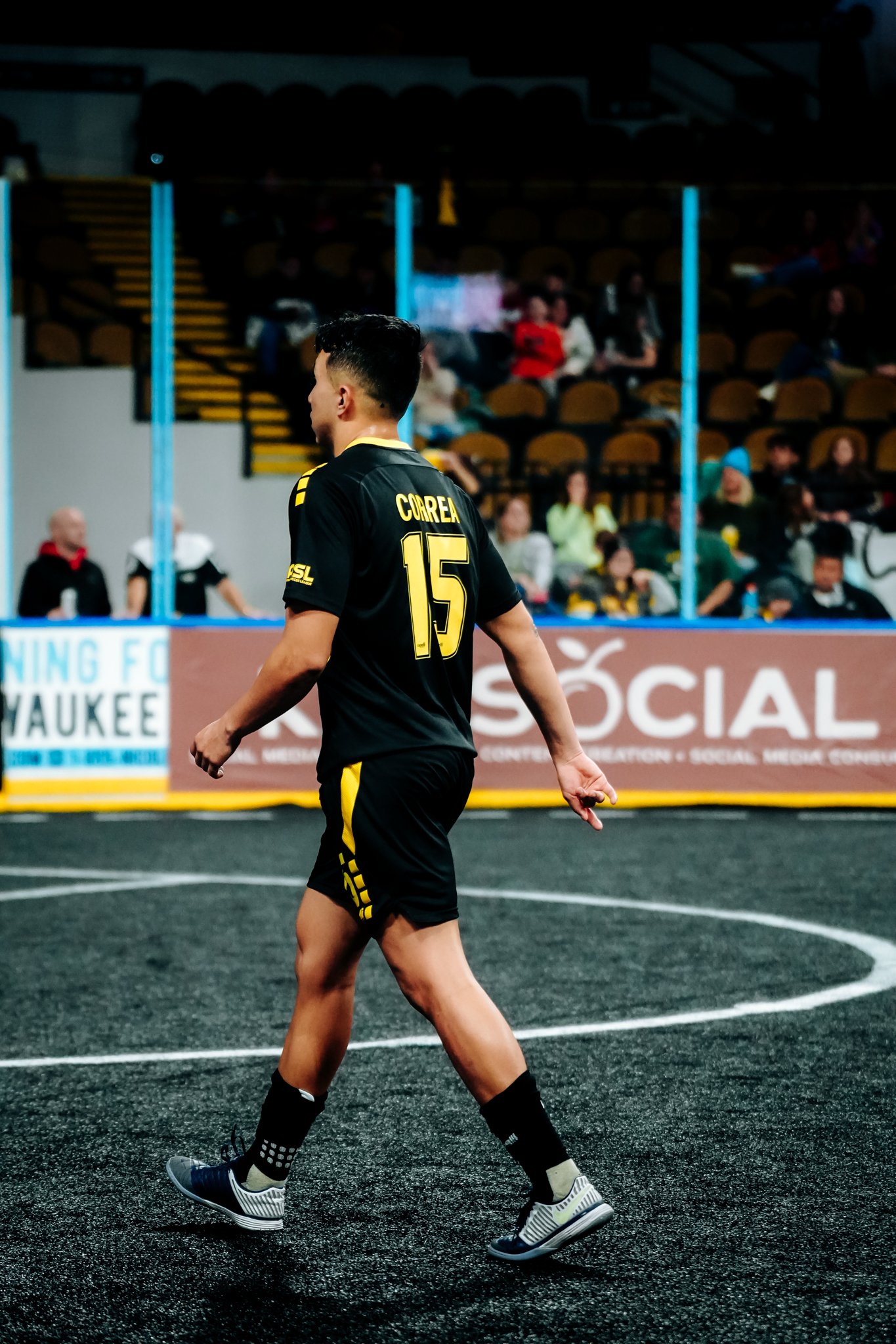 A soccer player wearing a black and gold jersey with the number 15 walks on an indoor field, with spectators seated in the background.