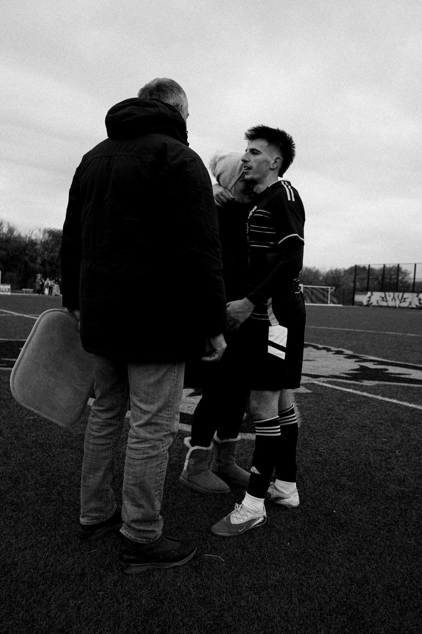 A soccer player in uniform having a conversation with a man in a jacket on the field, with a child on the player's shoulders.