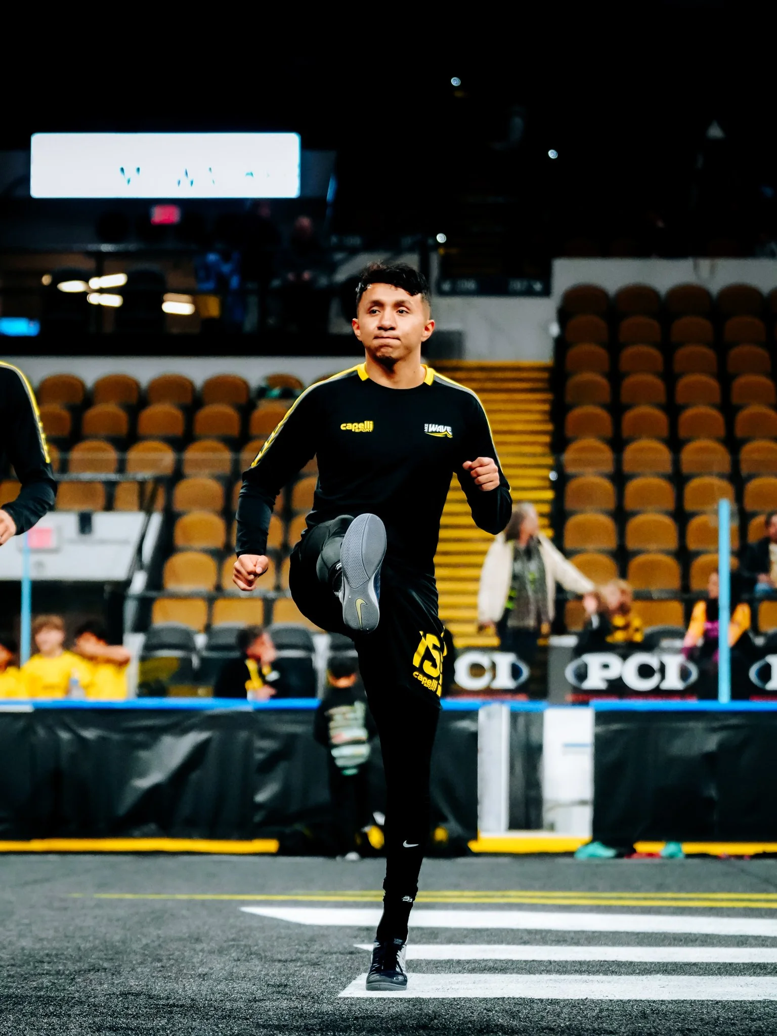 A male athlete dressed in black and yellow sportswear is warming up or running on a track or stadium floor. He appears focused, and the background shows empty seats and some people, possibly spectators or teammates, in an indoor sports arena.