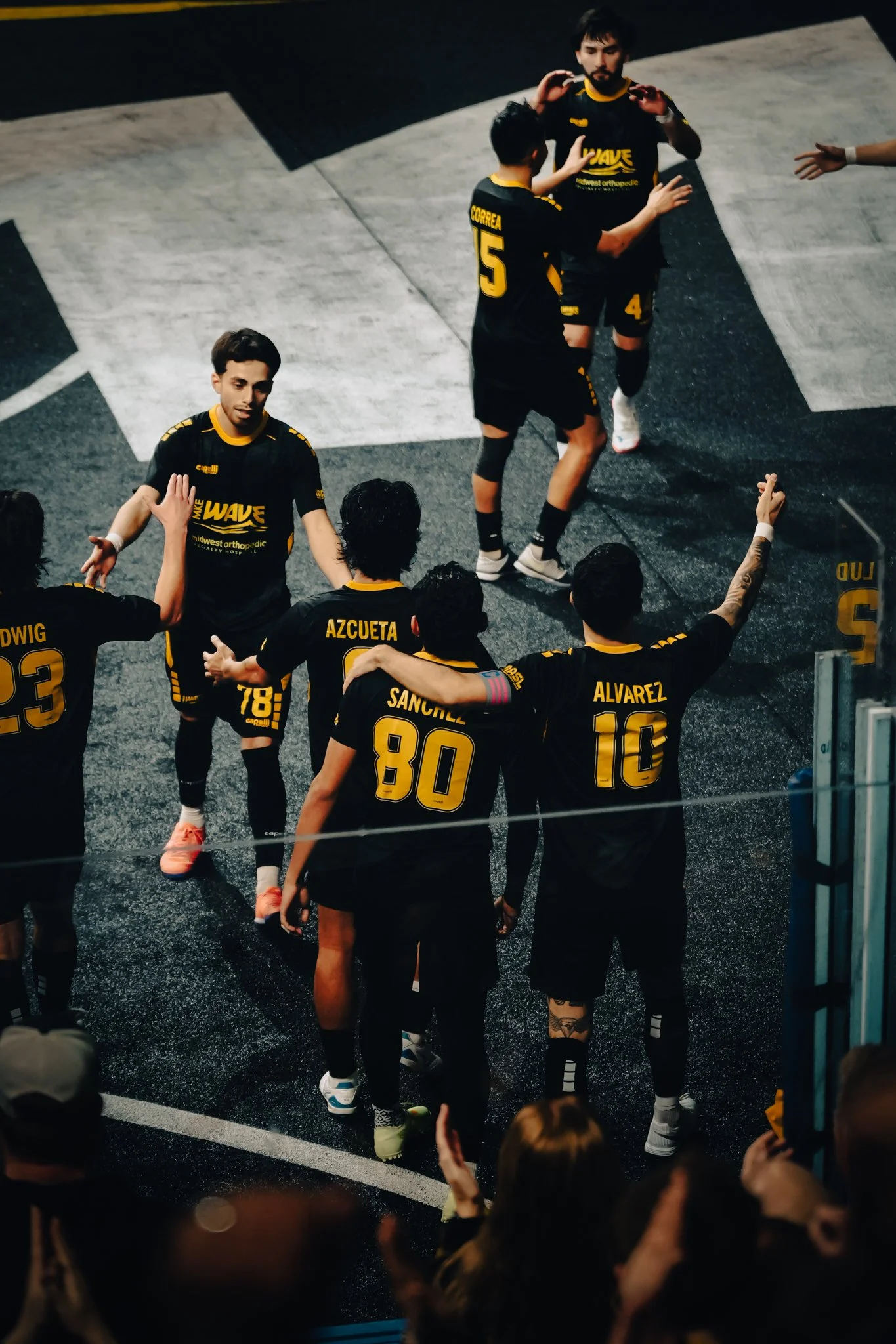 Group of volleyball players in black and yellow uniforms celebrating on the court, some giving high-fives.