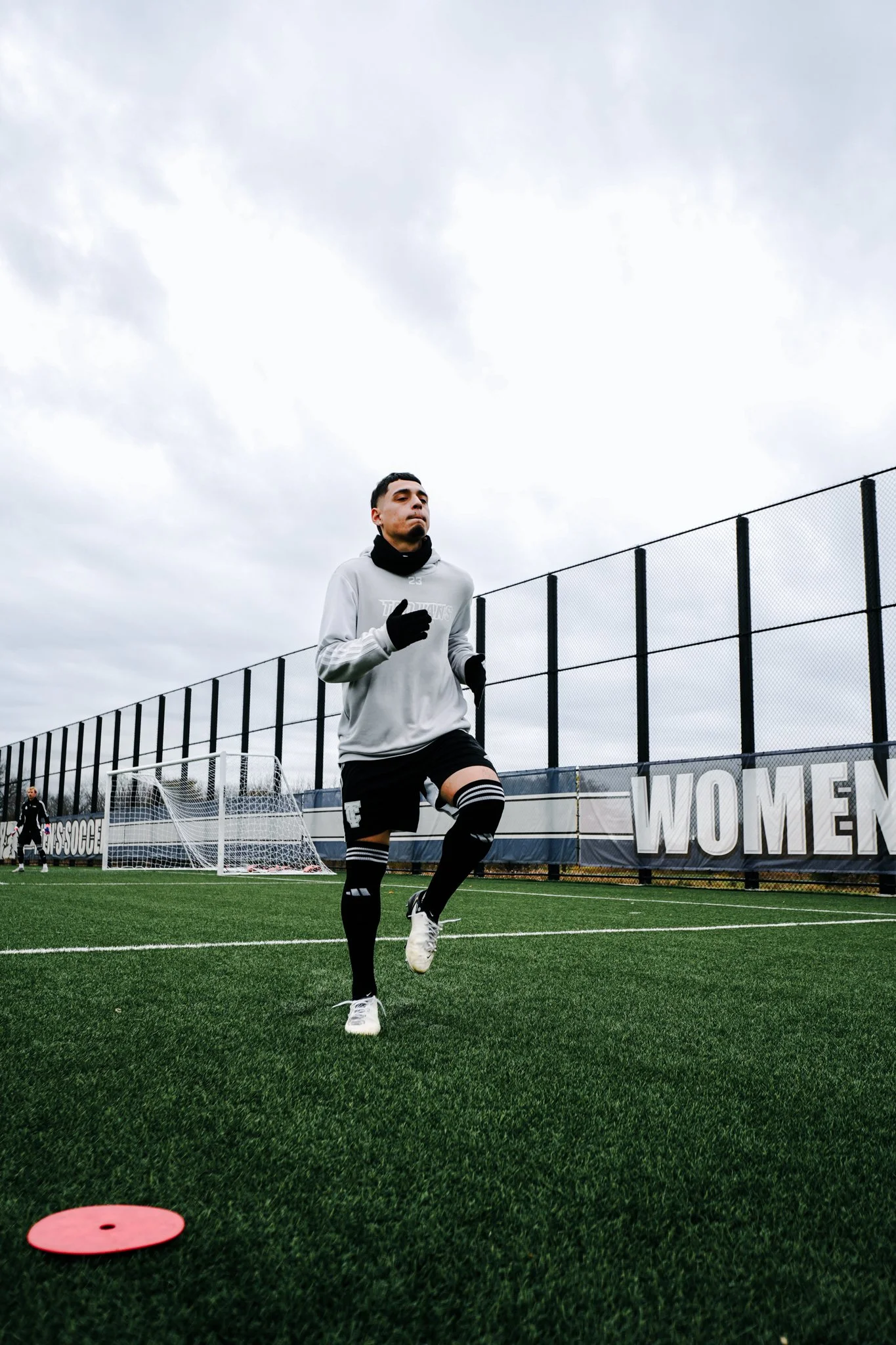 A male soccer player jogging on a soccer field, with a fence and goalposts in the background, on a cloudy day.