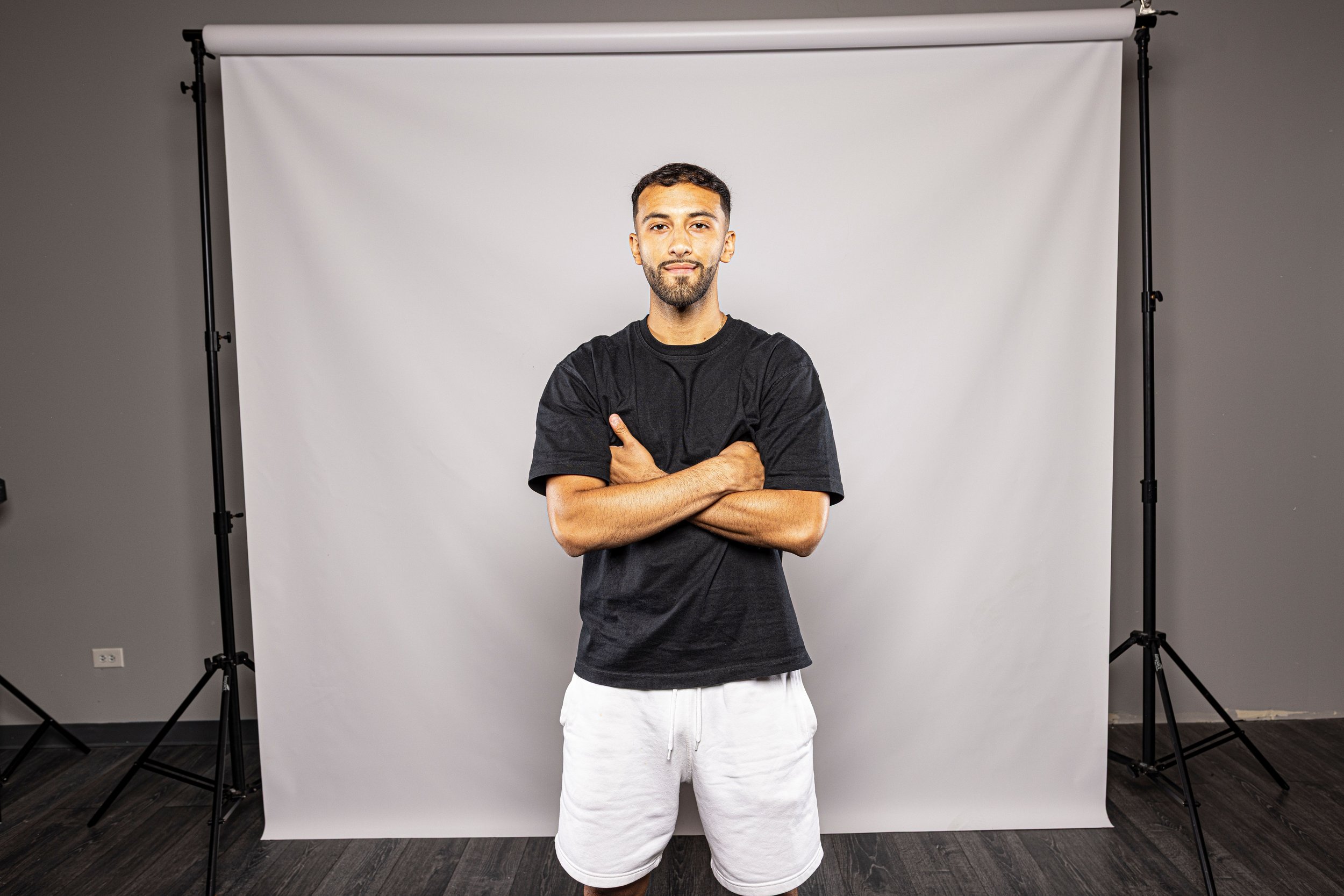 A young man stands with arms crossed in front of a white photography backdrop, wearing a black t-shirt and white shorts in a studio setting.