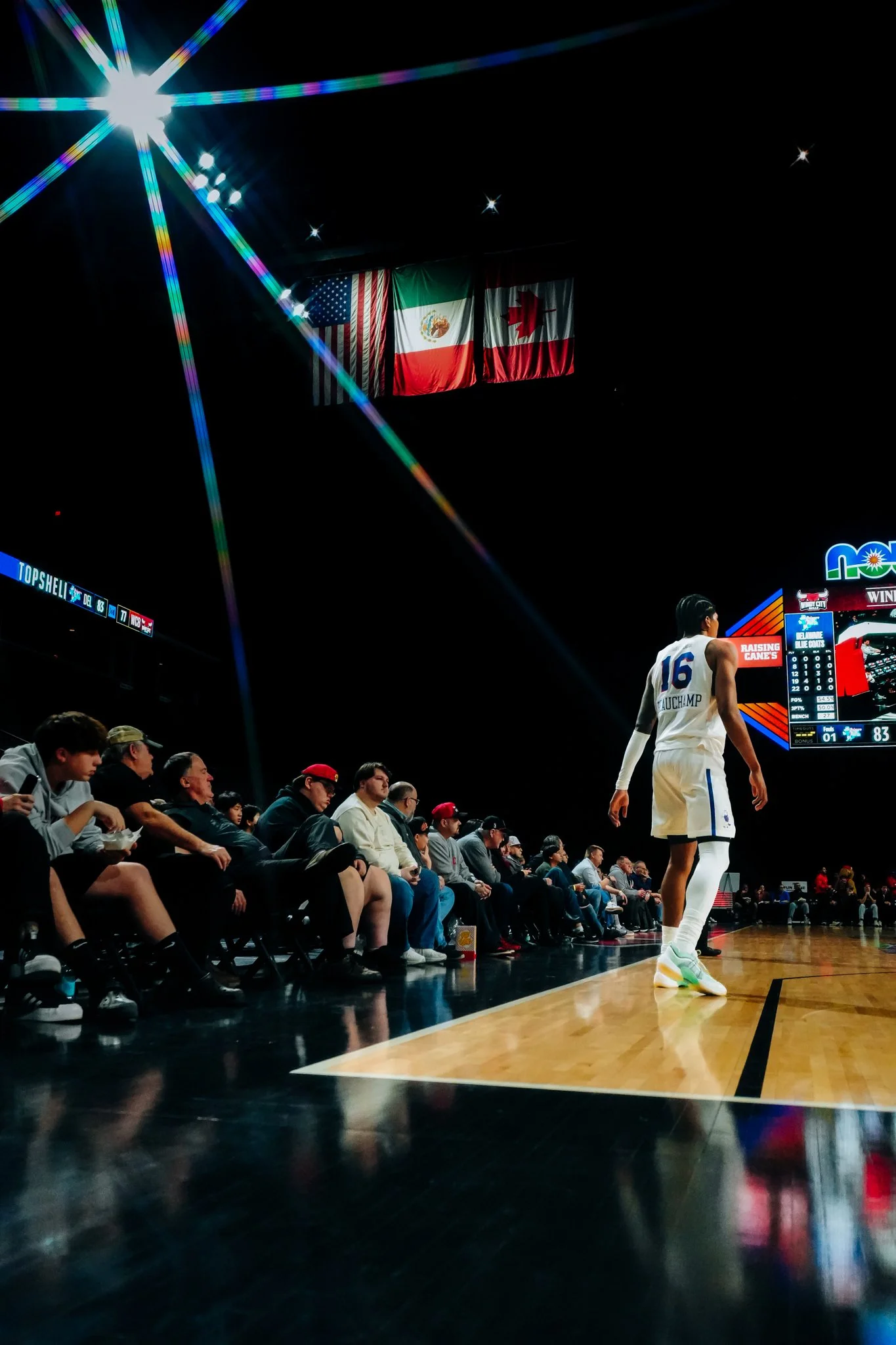 A basketball player wearing jersey number 16 with 'Tauchamp' on the back walking past seated spectators during a game, with flags of the United States, Mexico, and Canada above the court, and a scoreboard showing a score of 83-1.