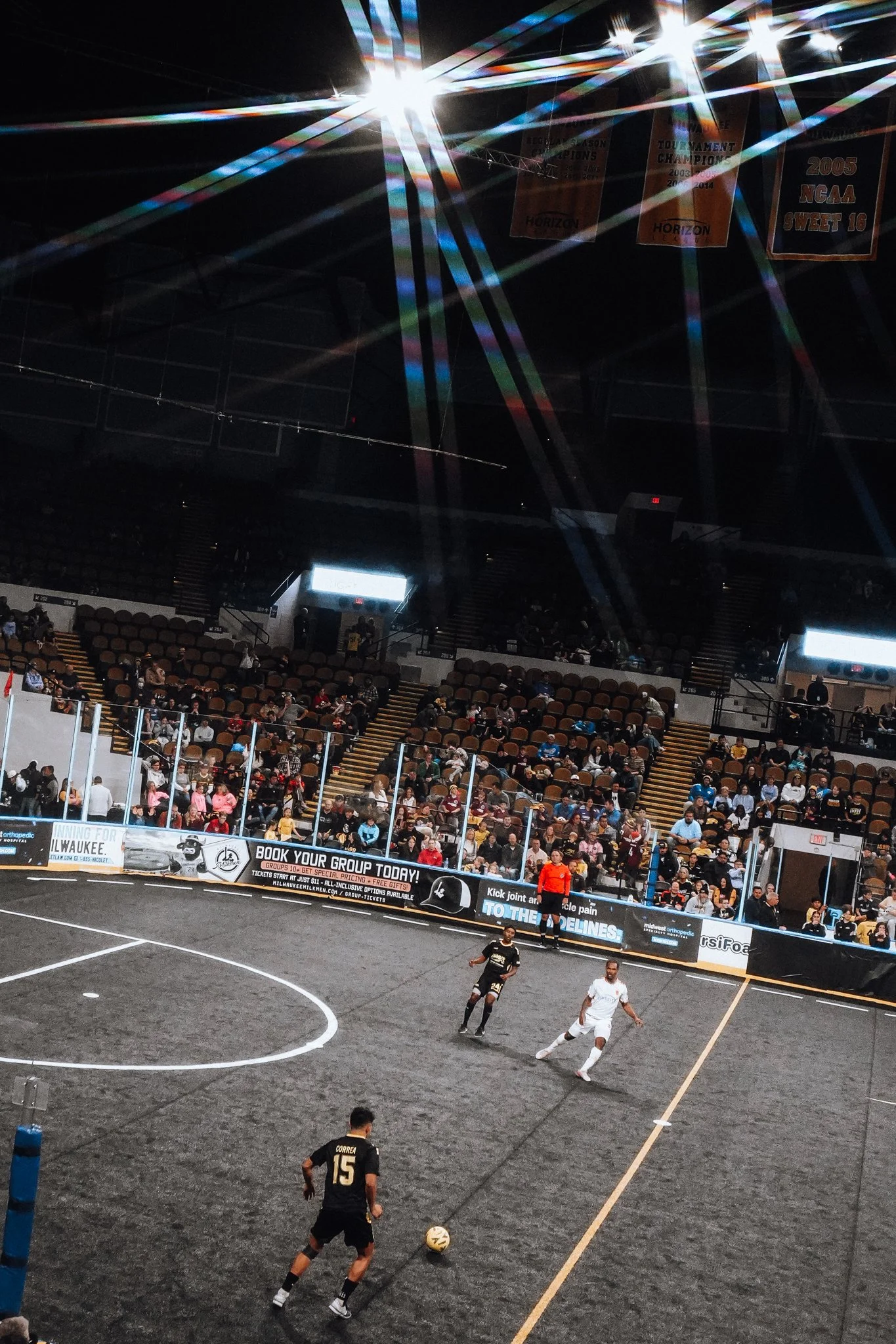 An indoor soccer match with players on a black turf field, surrounded by a crowd of spectators in the stands, under bright overhead lights with lens flares.