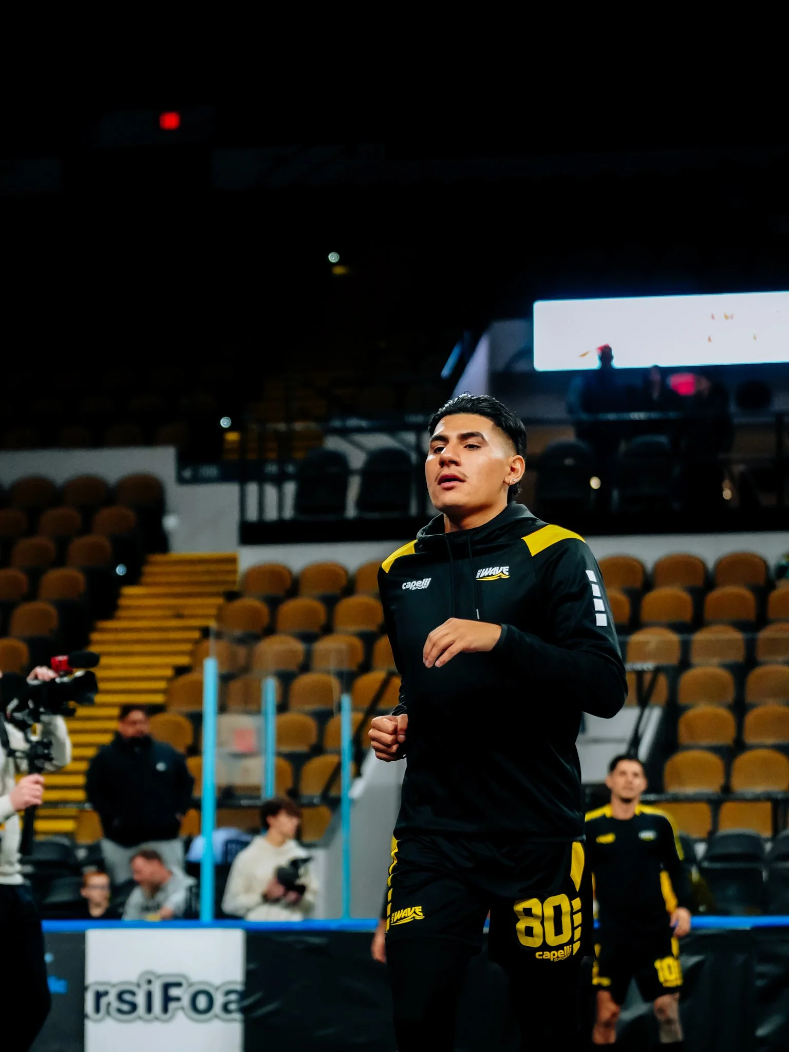 Young male athlete in black and yellow sportswear jogging indoors at a sports arena, with spectators and camera crew nearby.