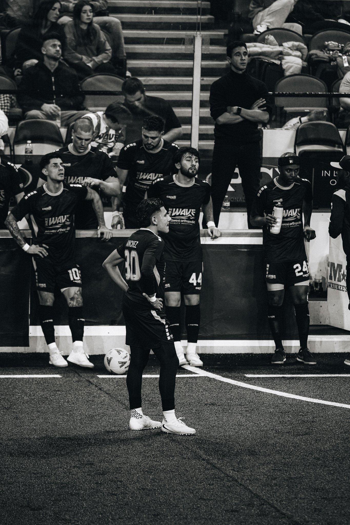 A group of soccer players in black uniforms standing and sitting near the sideline of a soccer field, with some spectators seated in the background.