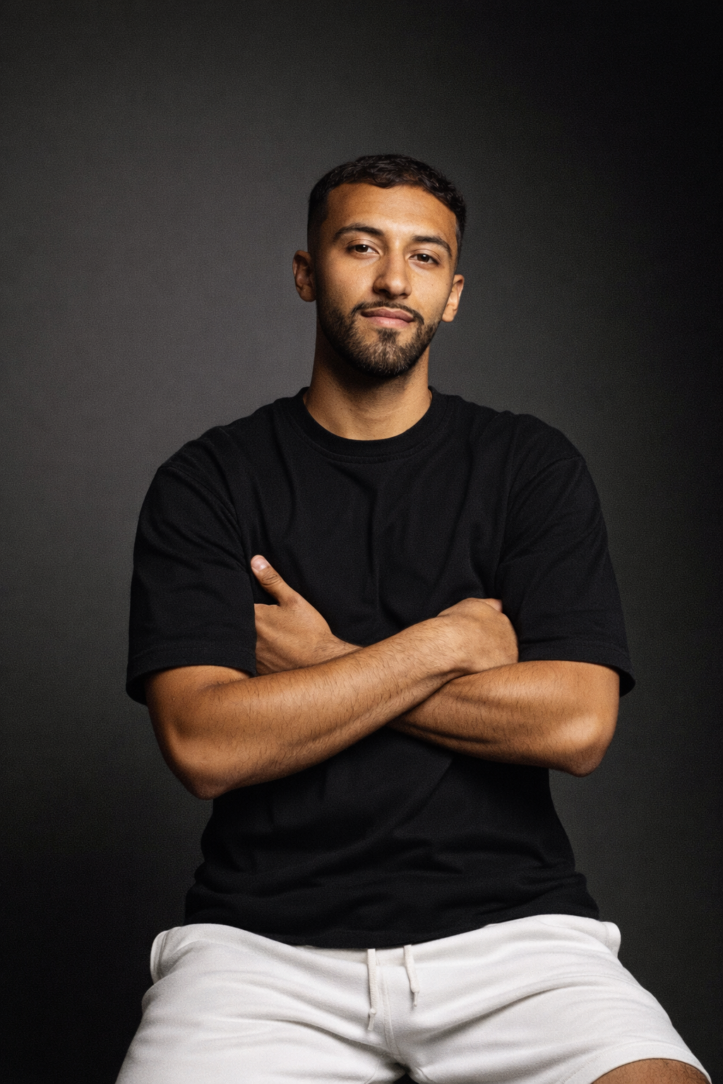A young man with a beard and short dark hair, wearing a black t-shirt and white shorts, sitting against a dark background with his arms crossed, looking at the camera with a slight smile.