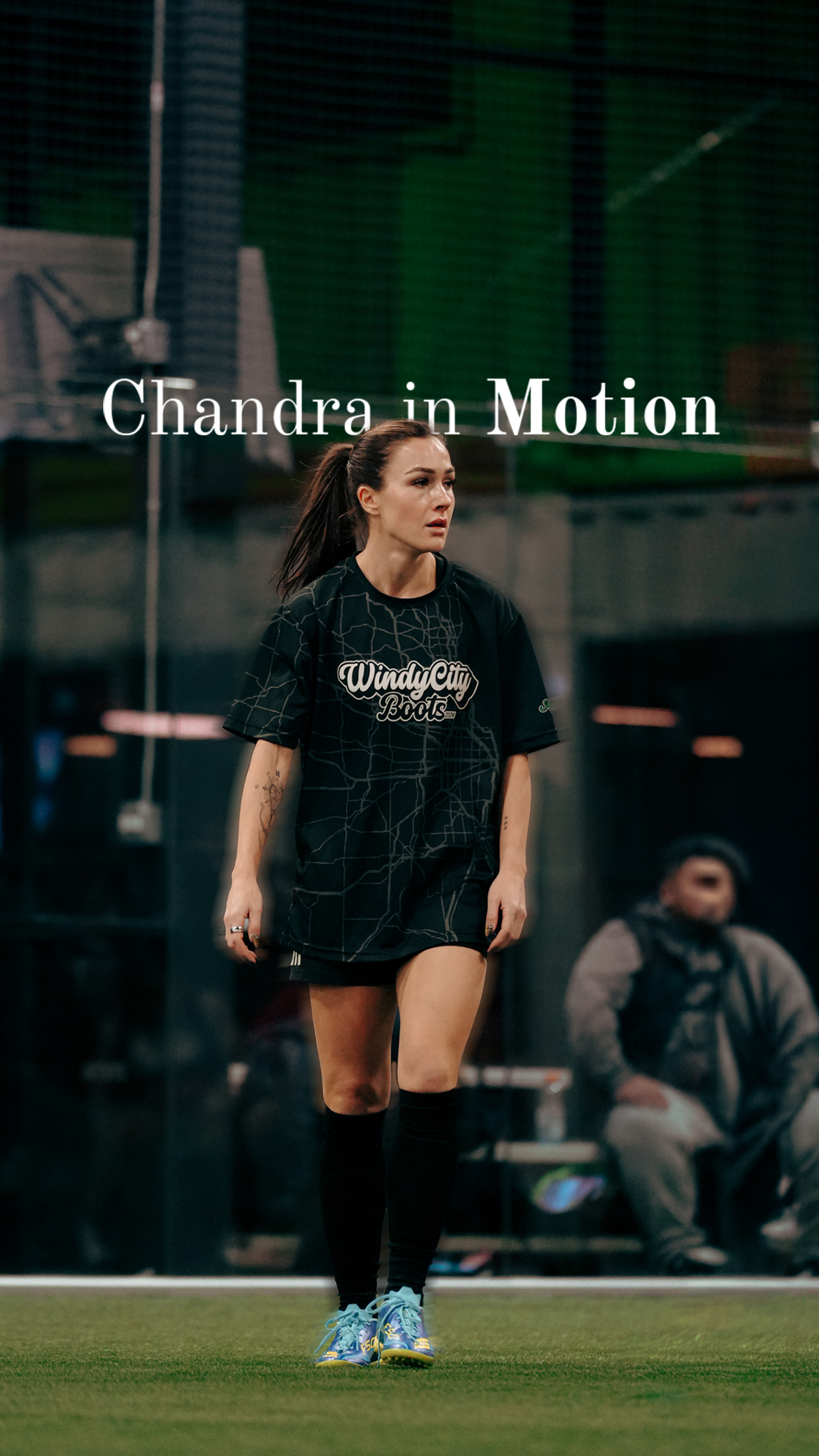 A woman in a black sports shirt and shorts walking on a sports field, with a man sitting in the background.