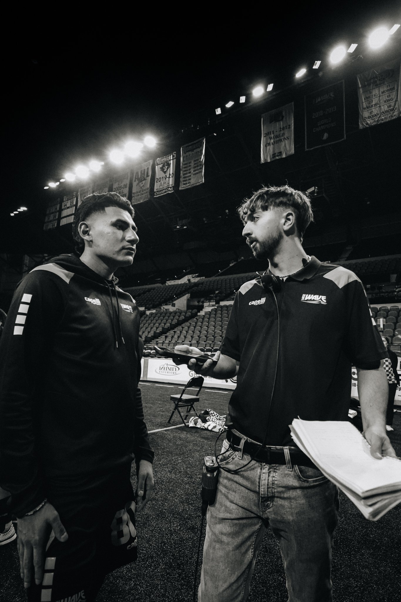 Two young men having a conversation in an indoor sports arena, one holding a microphone and a notepad, with banners hanging from the ceiling in the background.