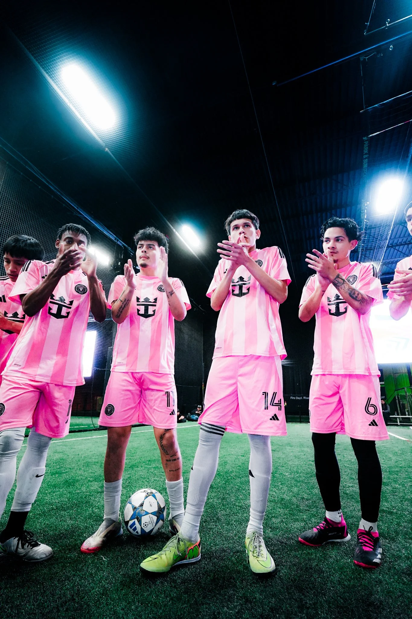 Soccer players in pink jerseys standing on an indoor field, clapping and looking forward, with one player resting a foot on a soccer ball.