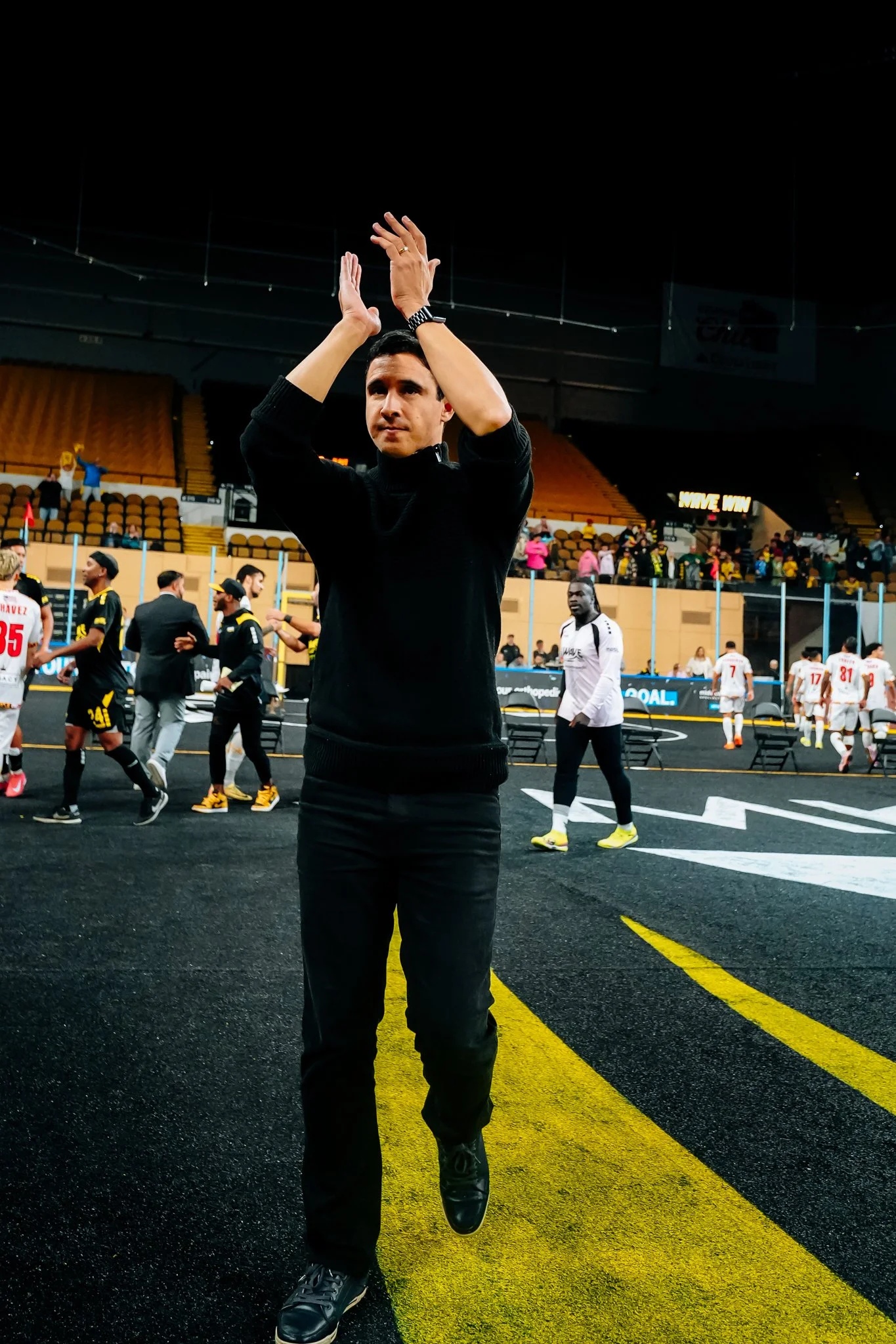 A man in a black sweater and pants claps his hands above his head on a sports field, with soccer players and officials in the background.