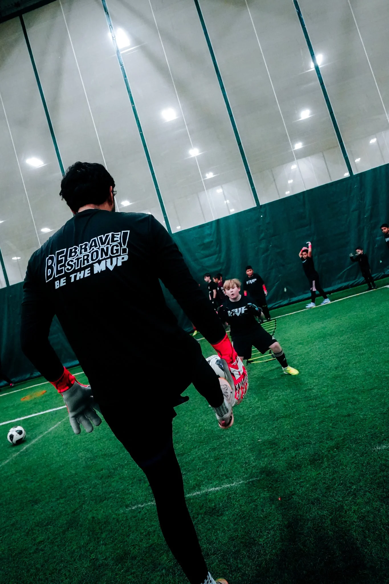 A coach instructs young children during a soccer practice inside an indoor facility with green turf and high ceiling.
