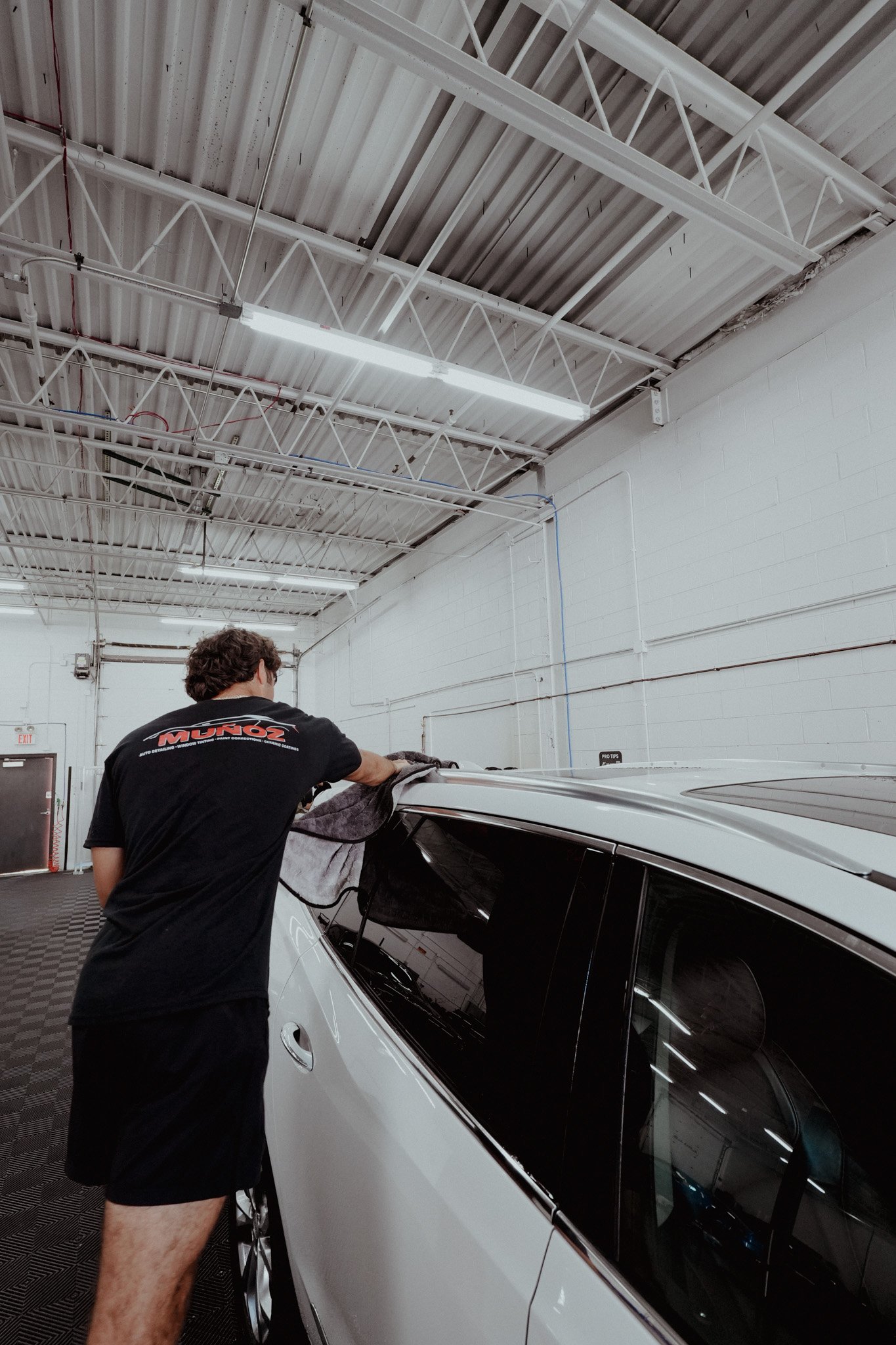 A person cleaning the side window of a white car inside a spacious, well-lit garage.