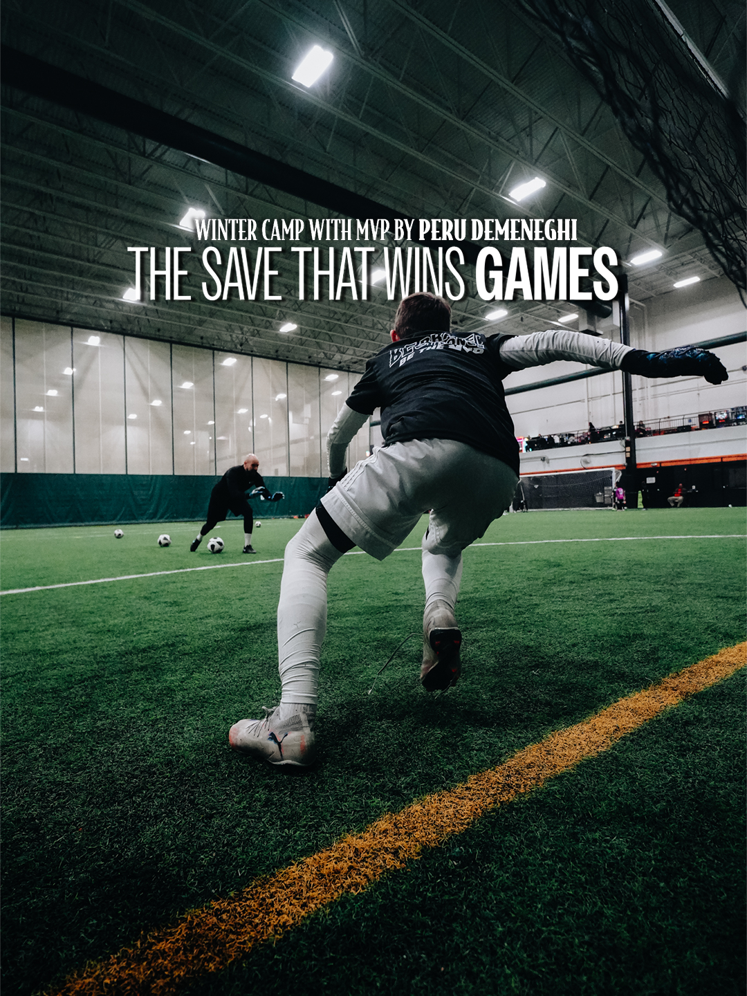 Indoor soccer field with two players practicing goalkeeping, one in the foreground wearing white and black, and a goalkeeper in the background near multiple soccer balls.
