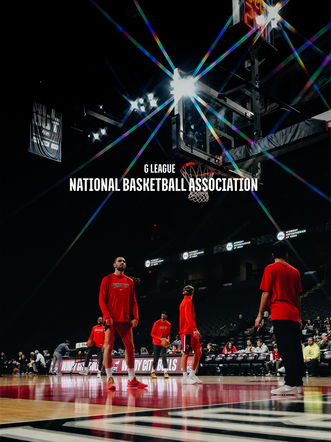 Basketball players warming up on the court at an NBA game, with the scoreboard and arena seating in the background, under bright lights and digital advertising screens.