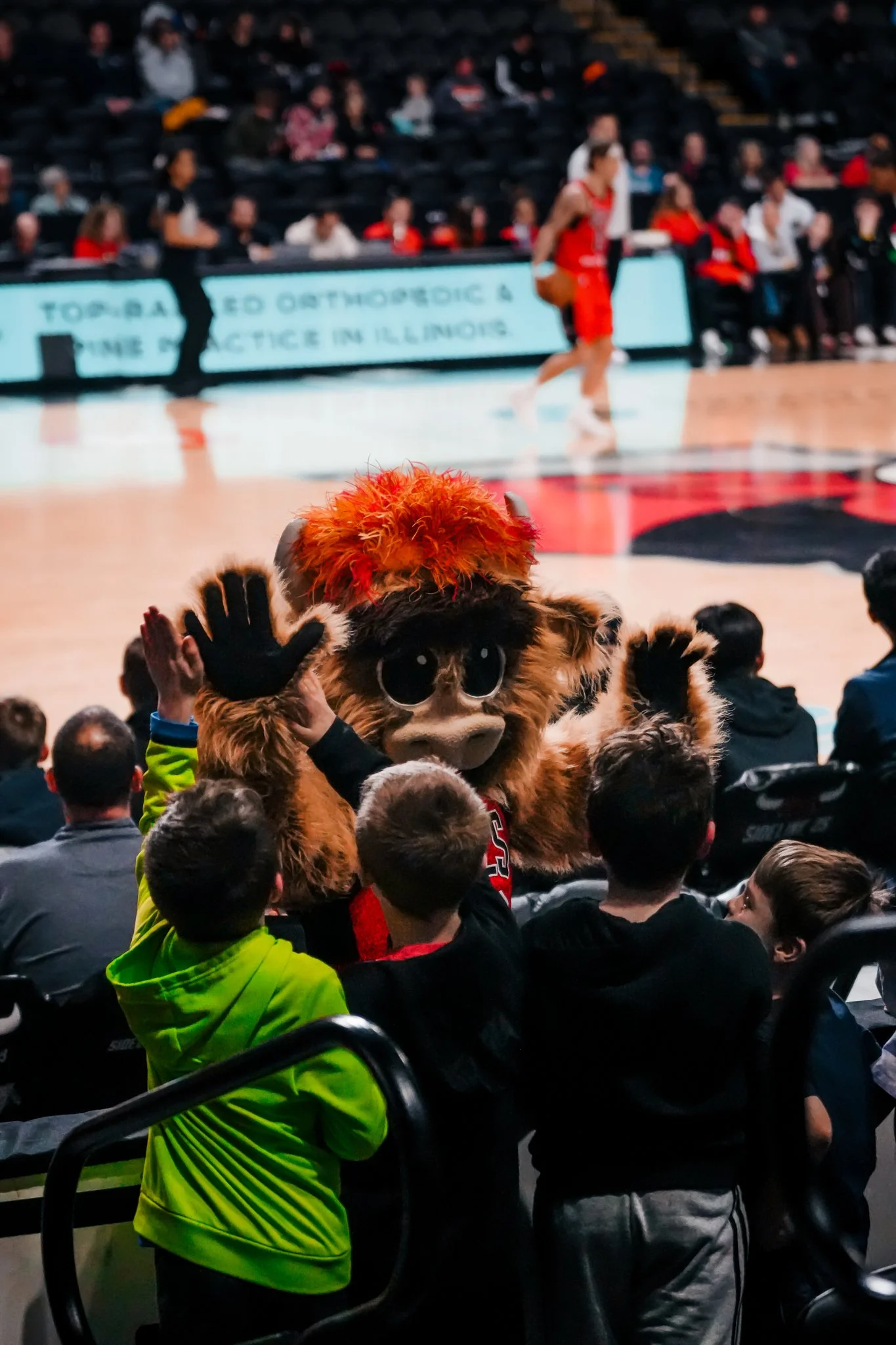 Basketball game in progress with fans watching, including a mascot in costume with large eyes, furry brown body, colorful hair, and high-fiving fans.