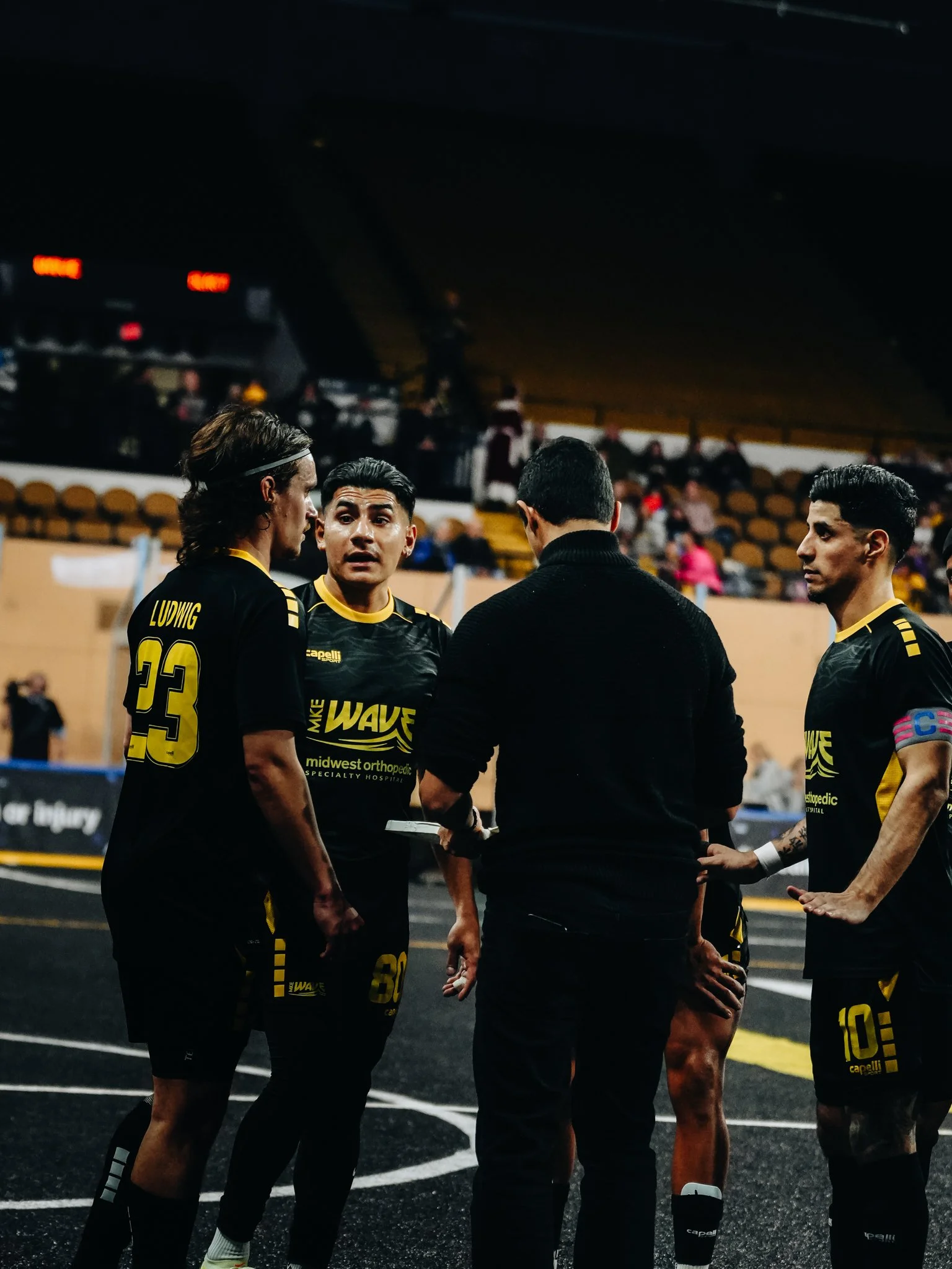 Four soccer players and a coach or referee in discussion during a game, with a mostly empty stands in the background.